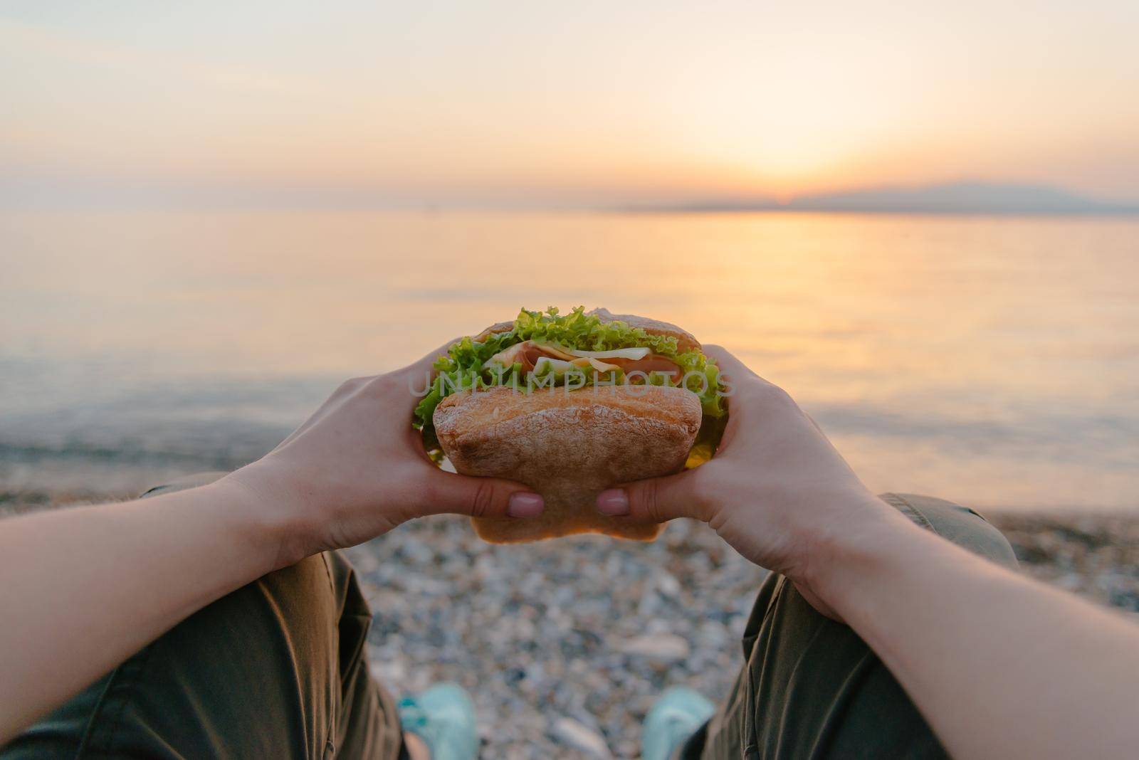 Young woman holding fresh tasty fast food burger sandwich and resting by the sea at summer sunset, point of view.