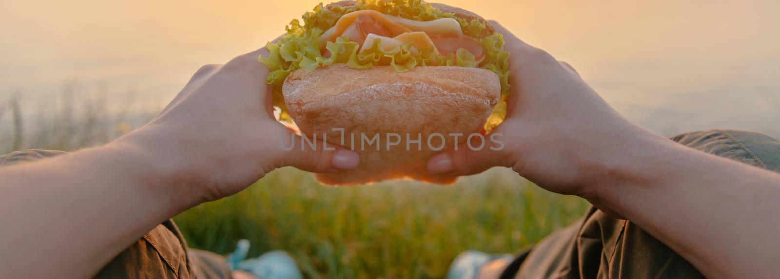 Unrecognizable young woman holding fast food fresh burger sandwich while resting on nature, point of view.