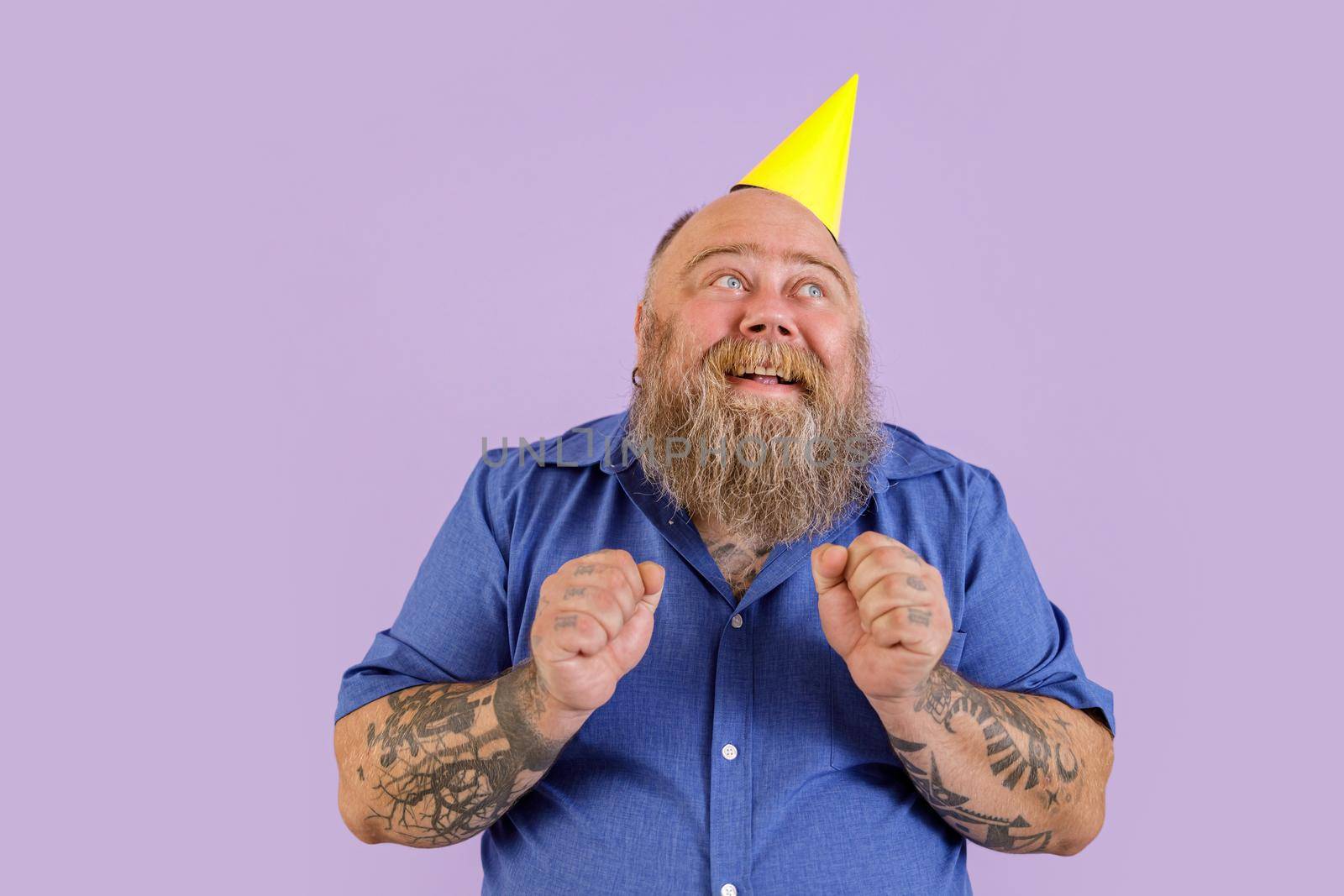 Hopeful middle aged bearded male person with overweight in party hat holds fists standing on purple background in studio