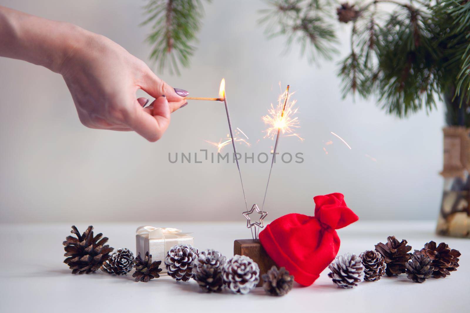 woman hand lighting festive sparkles near pine cones, christmas tree and red christmas hat. New year party burning sparkler closeup on white background. fireworks, shining fire flame. Christmas light.