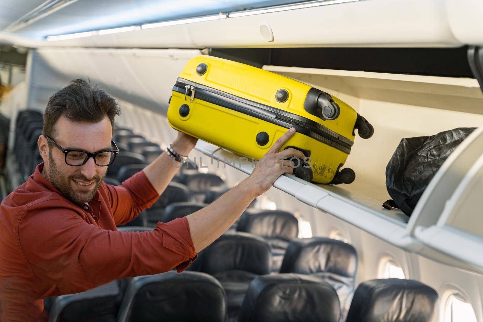 Smiling adult man with glasses putting a suitcase on a shelf in an airplane. Trip concept