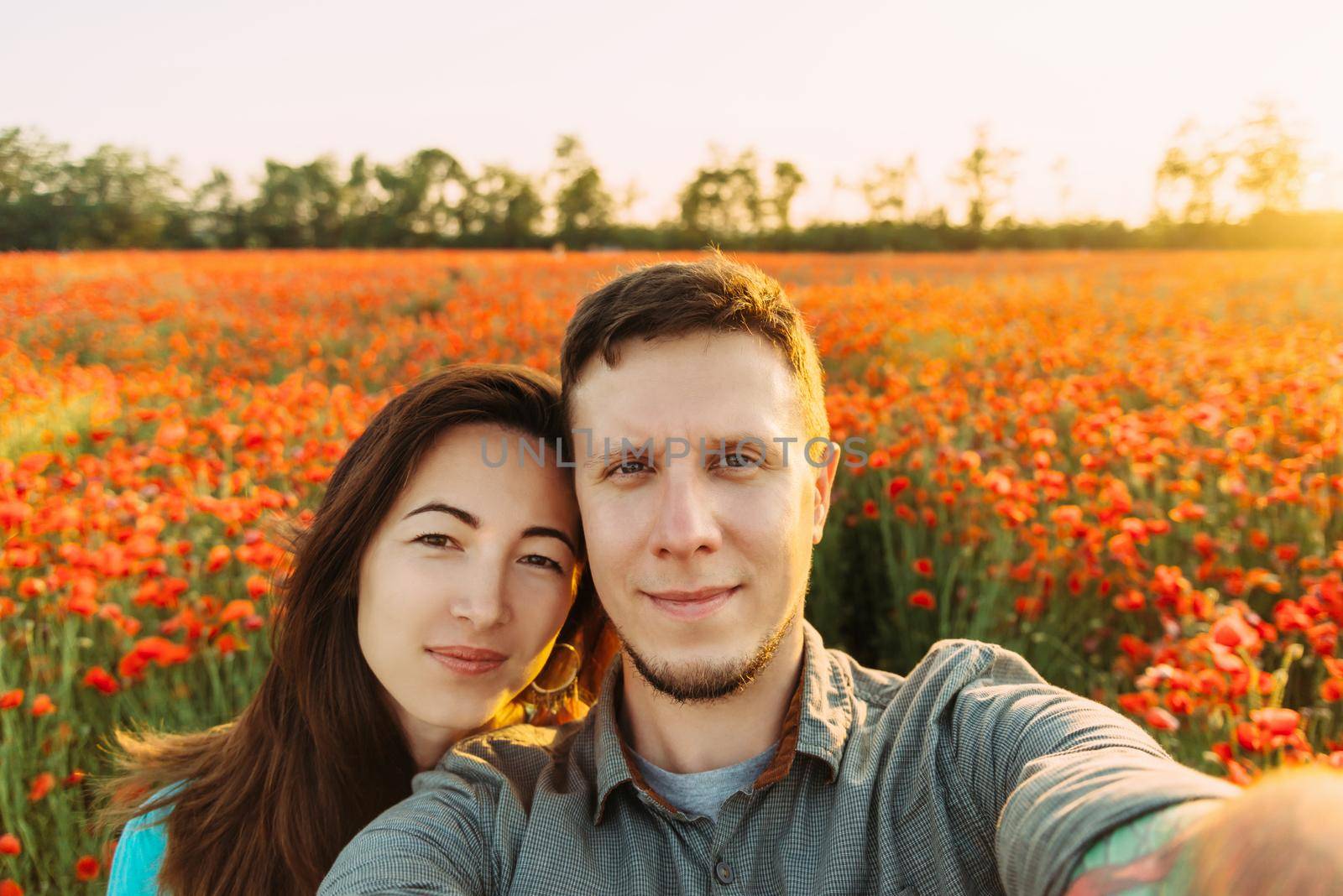 Smiling young loving couple taking photo selfie in red poppy flower meadow on sunny summer day, point of view.