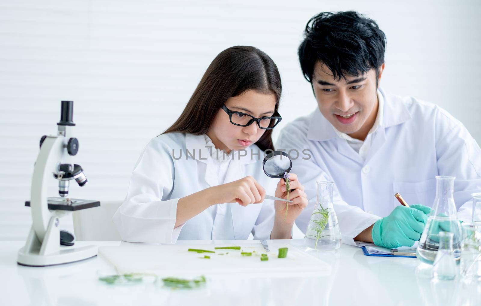 Little girl as young scientist use magnifying glass to analyze plant tissue and discuss with her teacher who sit near and consult the experiment also record the data in laboratory or classroom.