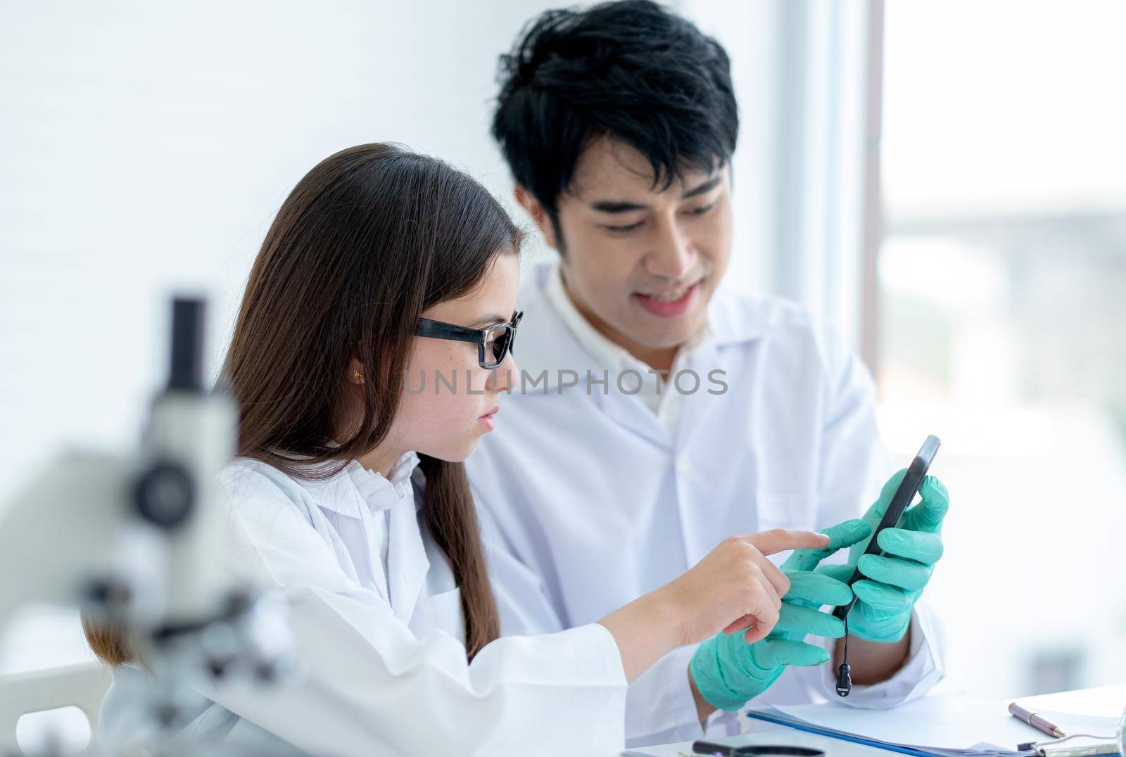 Young Caucasian scientist child girl point to mobile phone that her Asian teacher explain about experimental analysis in laboratory or classroom.