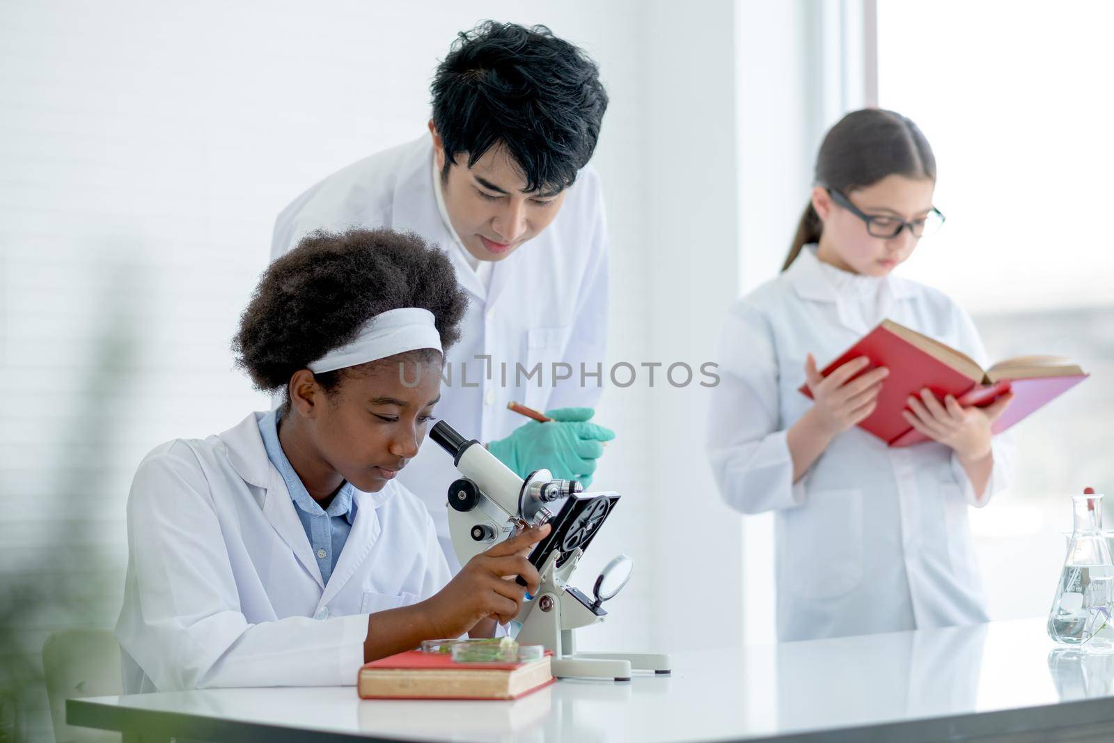 Asian teacher help and guide to African American young scientist child girl use microscope to analysis about plant tissue in laboratory or classroom while other student read some books.
