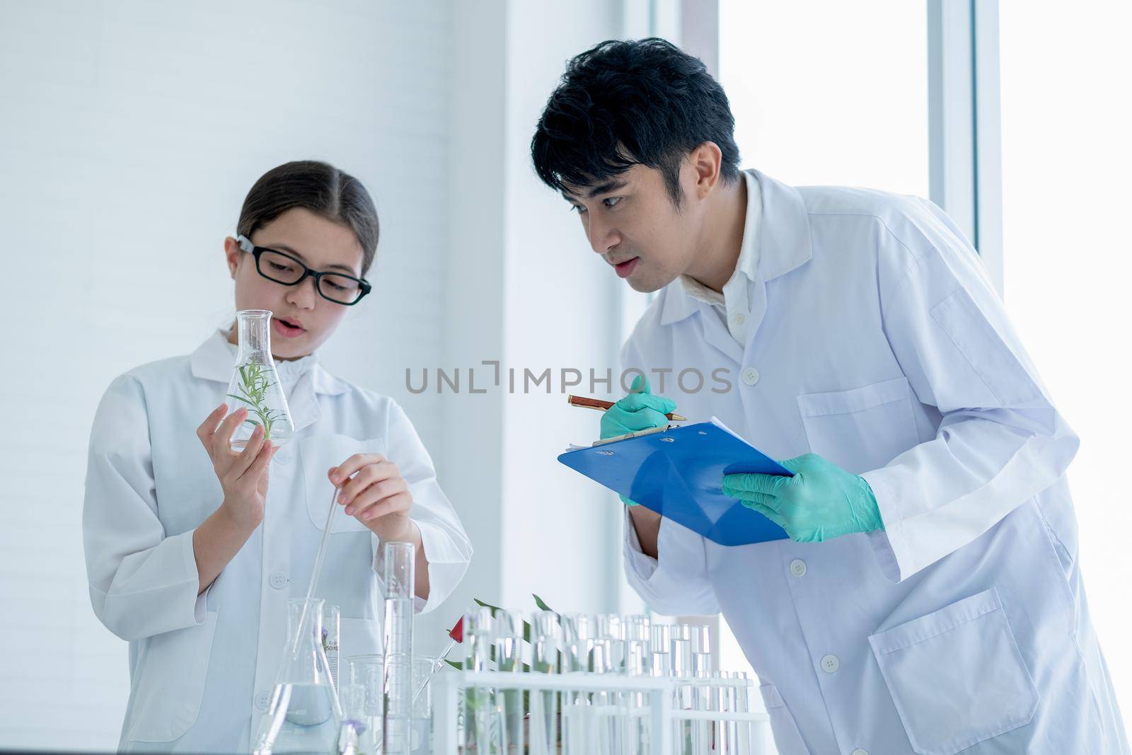 Young Caucasian scientist child girl hold flask with plant tissue and explain to her Asian teacher with record some data during do experiment in laboratory or classroom.