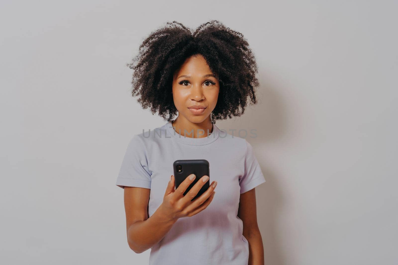 Studio portrait of young african american female with curly hair using mobile phone and looking at camera, mixed race woman with modern smartphone in hands isolated on light background