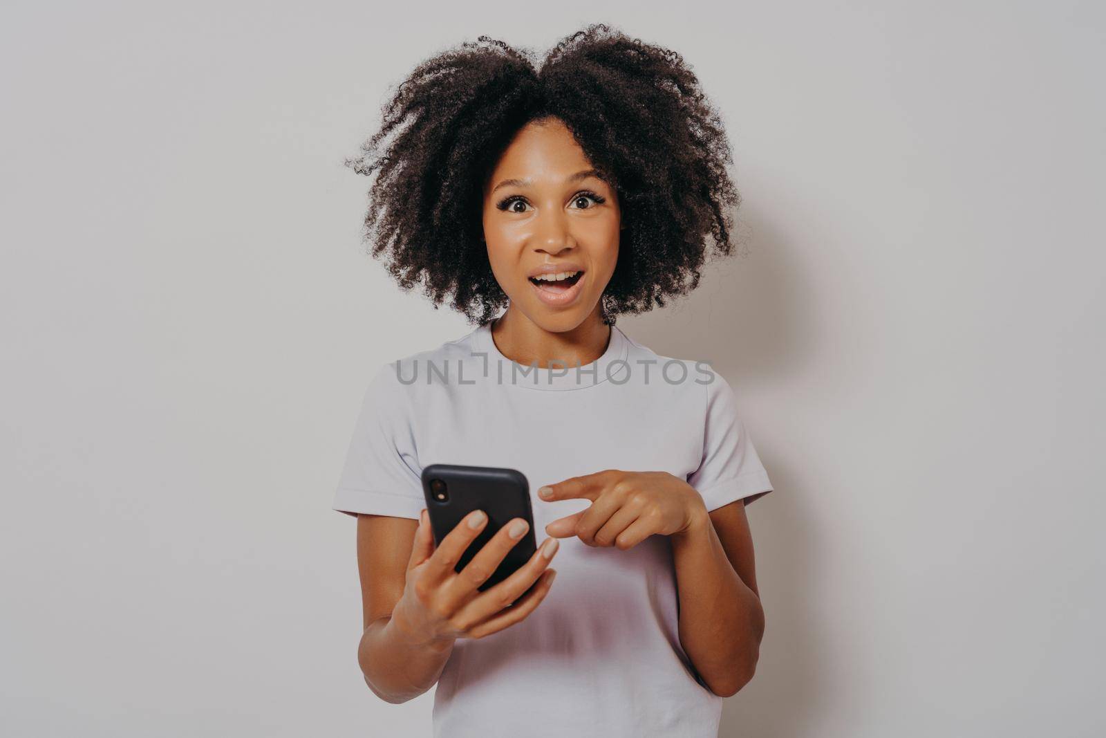 Excited happy dark skinned lady with curly hair holding mobile phone and reading good news notification, enjoying chatting with friends or doing online shopping, isolated on white studio background