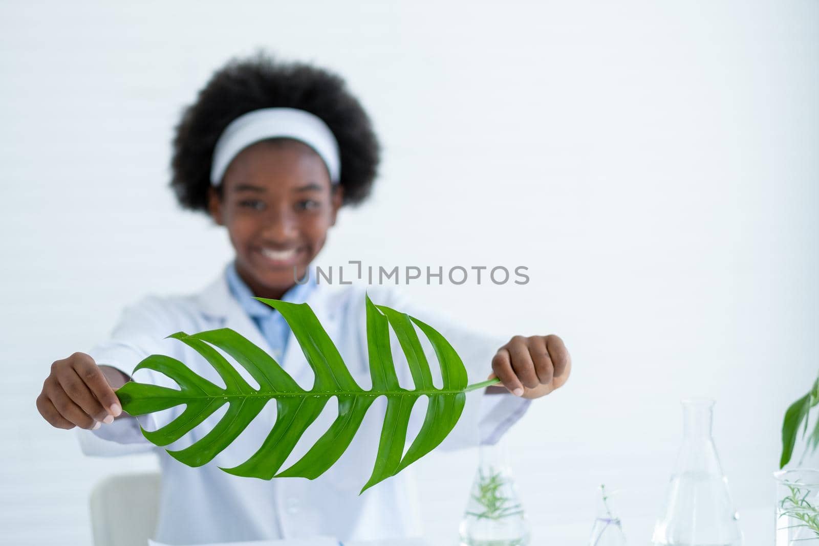 Main focus on green leaf is held by young American African scientist in laboratory or classroom.