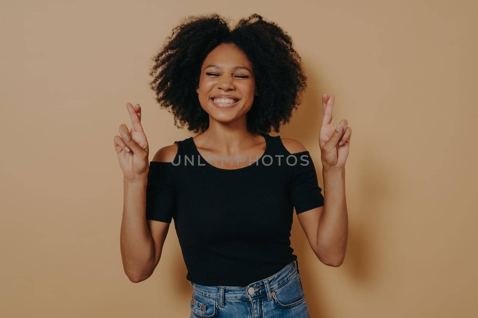 Cute lovely african american woman posing in casual clothes while gesturing fingers crossed sign, smiling with hope and eyes closed. Luck, superstitious and body language concept