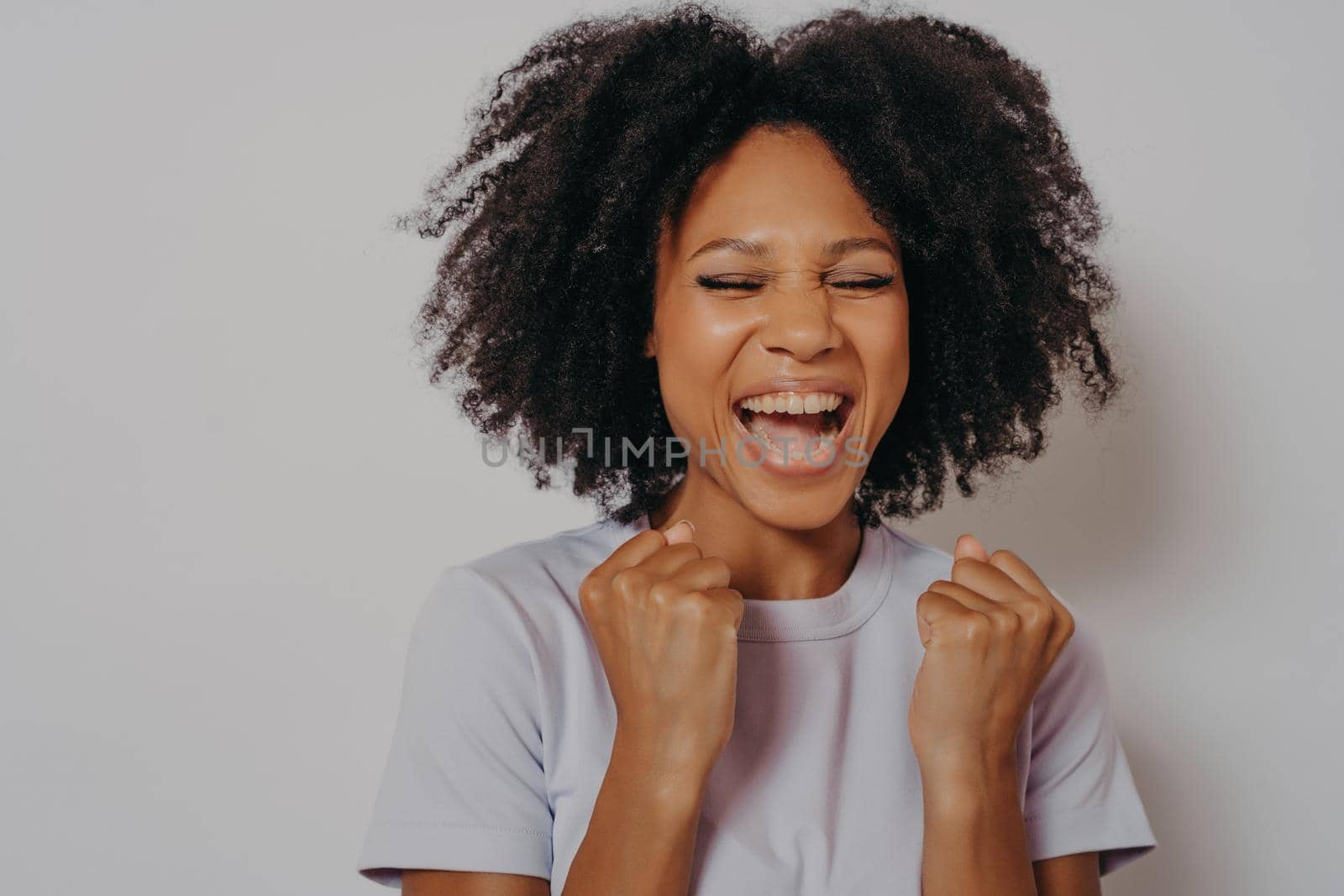 Excited cheerful dark skinned girl clenches fists while closed her eyes exclaims with victory or willing to win, excited female dressed in casual wear shouts yeah, isolated on white