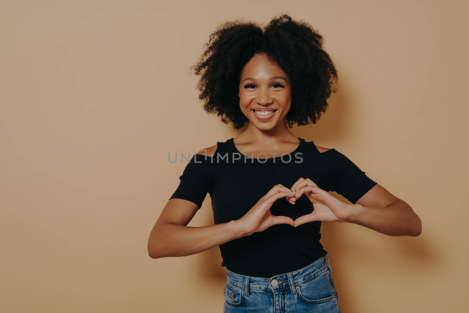 Close up photo of beautiful amazing dark skinned lady showing with hands sweetheart symbol, love message gesture, wears casual black t-shirt isolated over dark beige studio background with copy space