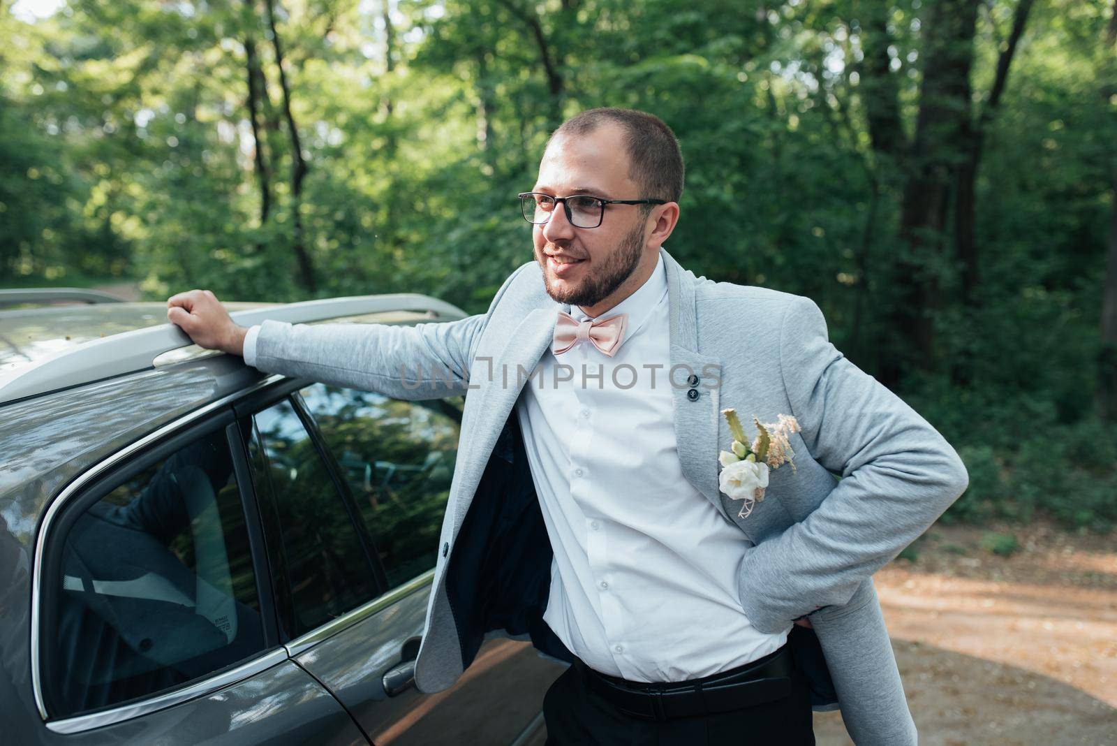 The groom with a beard in a gray jacket and glasses stands leaning on the car by lunarts