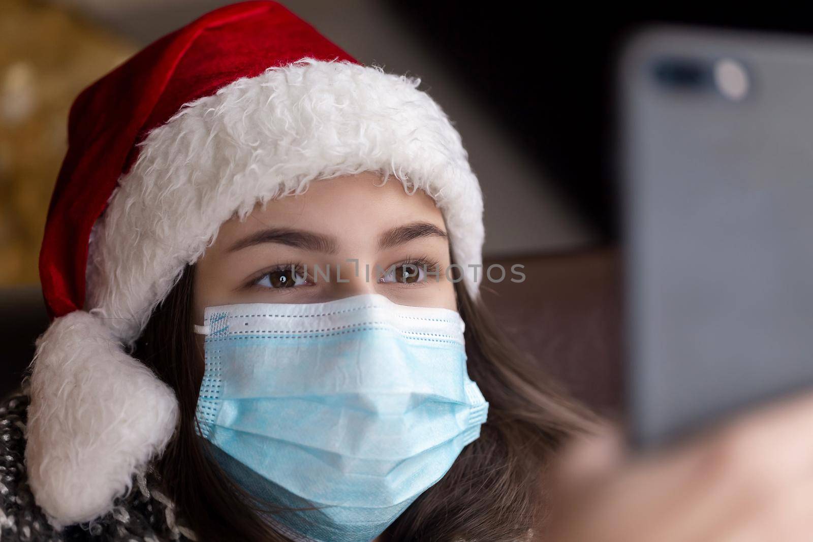 Christmas online greetings. A young girl in a santa claus hat and a medical mask talks using smartphone for video call friends and parents. Christmas during the coronavirus