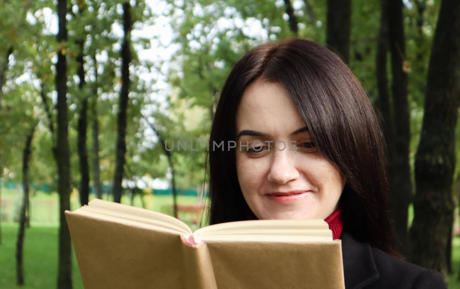 Smiling brunette woman reading a book in the park. Portrait of a happy charming Caucasian woman outdoors. by Roshchyn