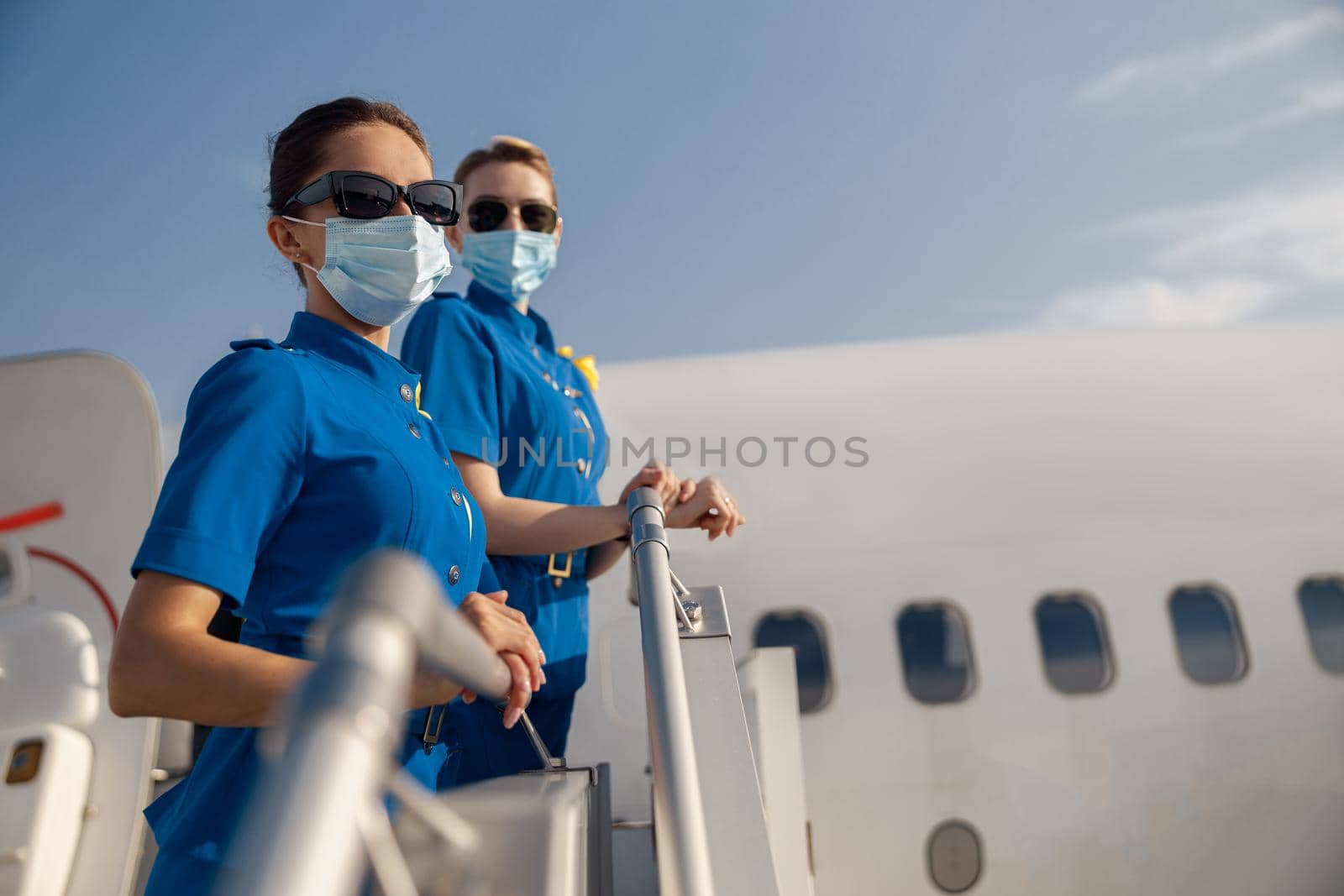 Two young air stewardesses in blue uniform, sunglasses and protective face masks looking at camera, standing on airstair on a daytime. Aircrew, occupation, covid19 concept