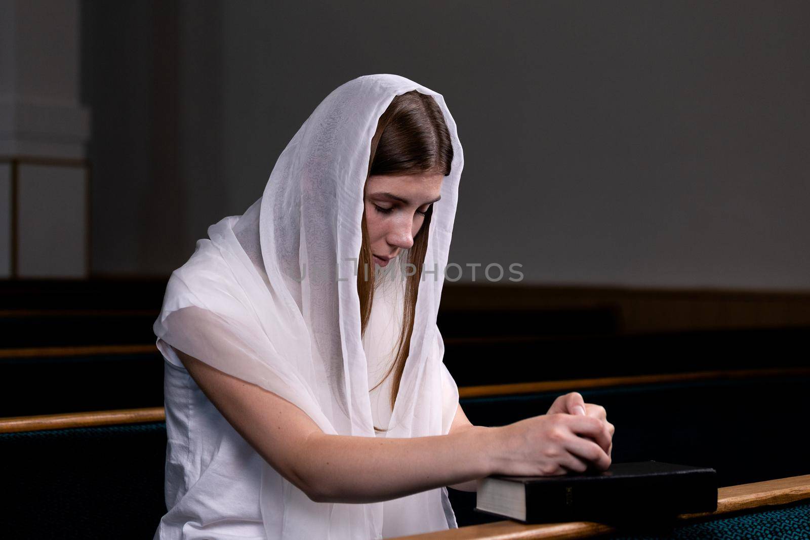 A young modest girl with a handkerchief on her head and a bible in her hands is sitting in church and praying. The concept of religion, prayer, worship by lunarts