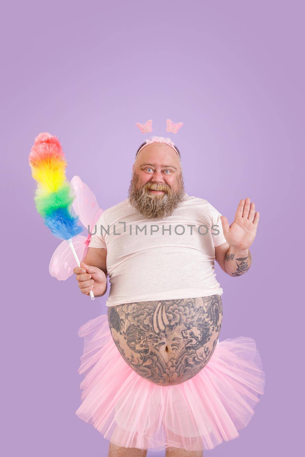 Cute bearded plus size person in carnival fairy suit with pink wings holds colorful dust brush on purple background in studio