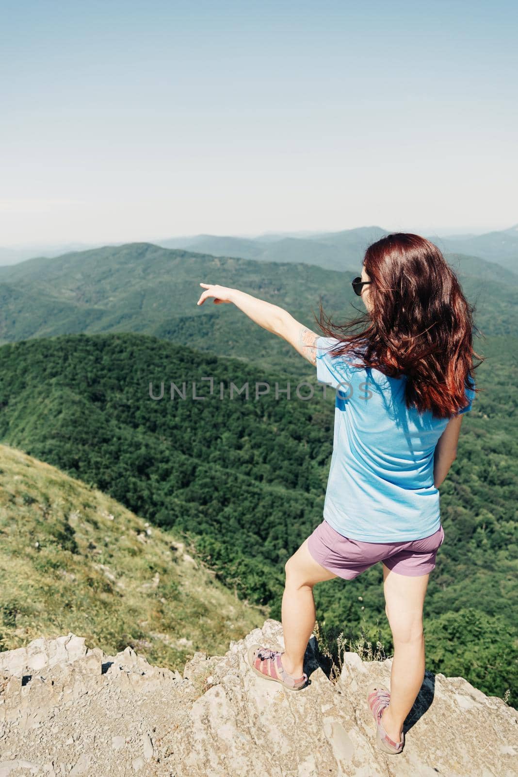 Sporty young woman standing on top of cliff and pointing at mountains in summer outdoor.