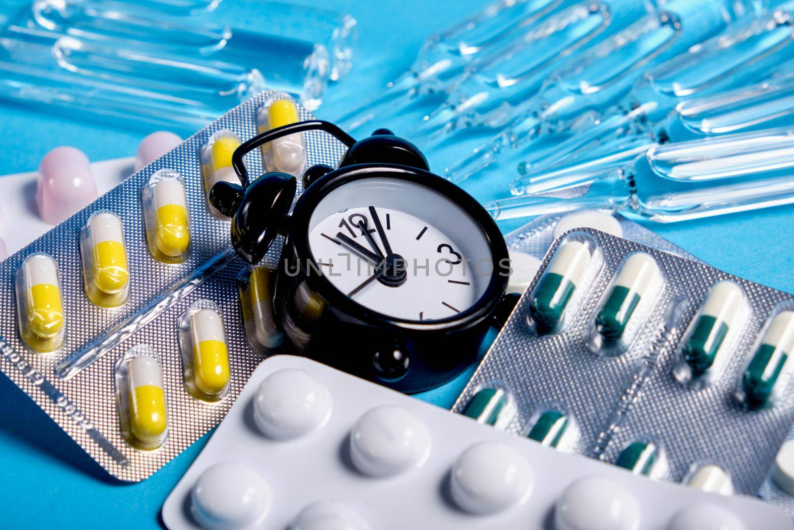 small black alarm clock, ampoules and packages of different pills on a blue background, copy space