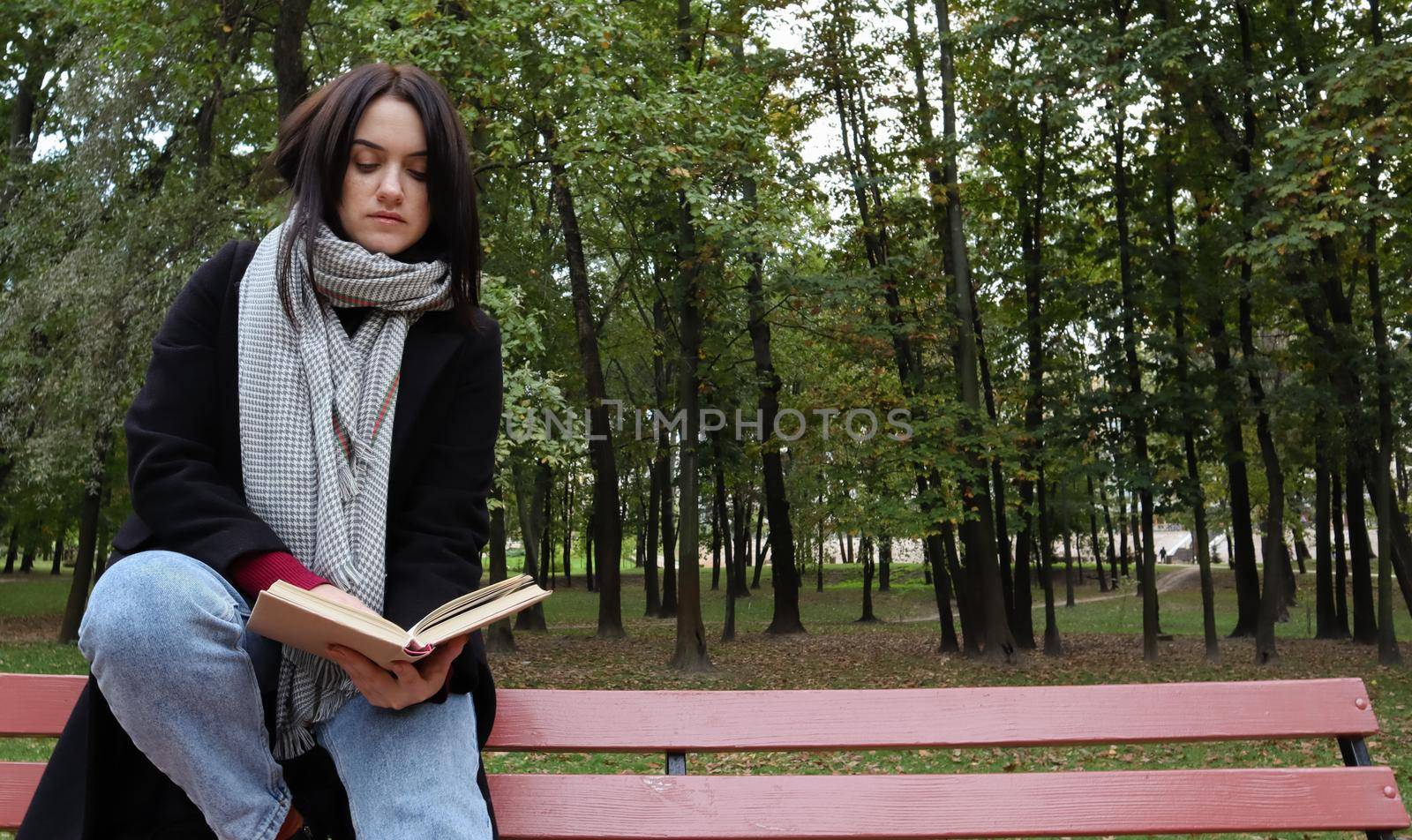 Young woman in jeans, coat and scarf, on a park bench. A woman is reading a book and drinking coffee or other hot drink outdoors alone. by Roshchyn