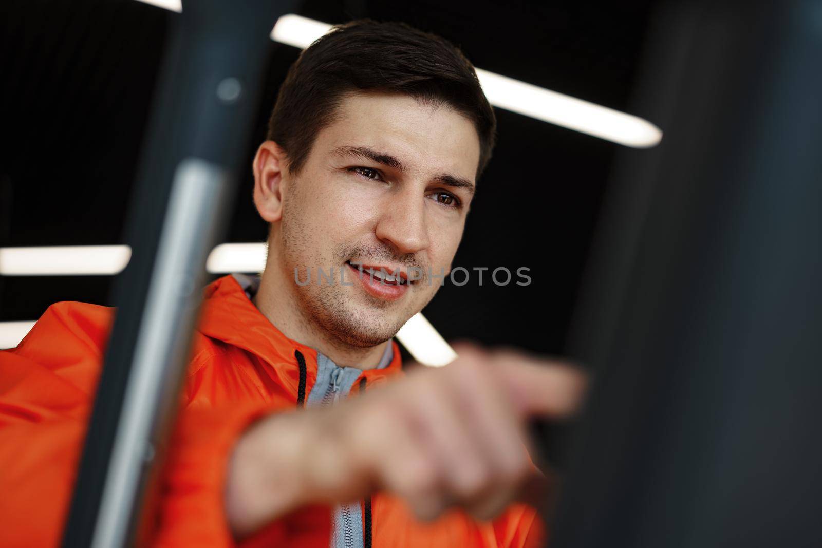 Portrait of a young man in orange windbreaker workout on a fitness machine at a gym.