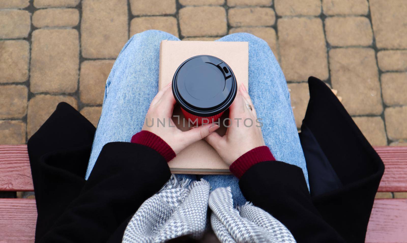 Young woman in jeans, coat and scarf, on a park bench. A woman is reading a book and drinking coffee or other hot drink outdoors alone. Close-up. View from above. The concept of honor, leisure. by Roshchyn