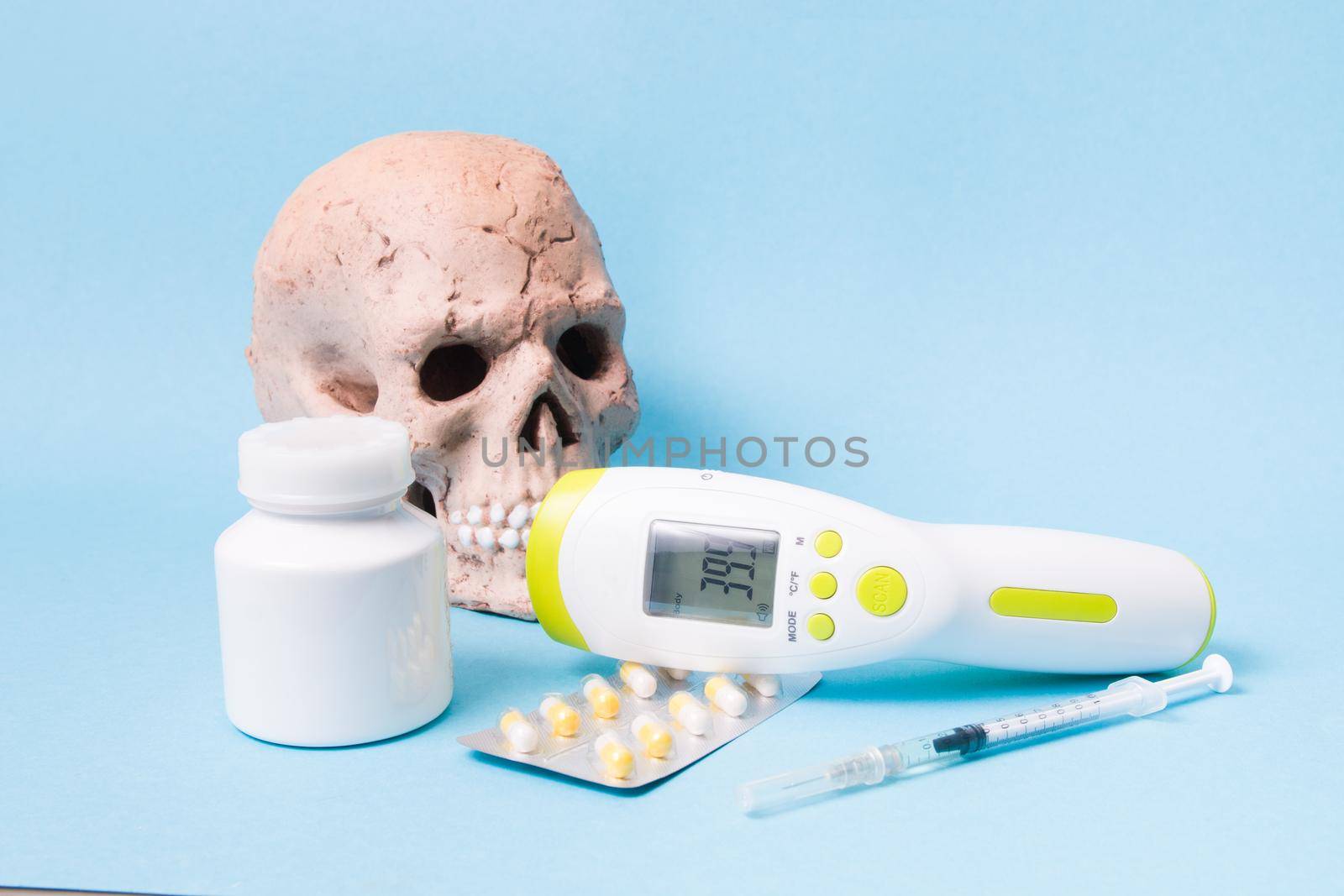 pills, drugs, syringe, infrared non-contact thermometer and ceramic skull in a protective medical face mask on a blue background, copy space, high temperature concept