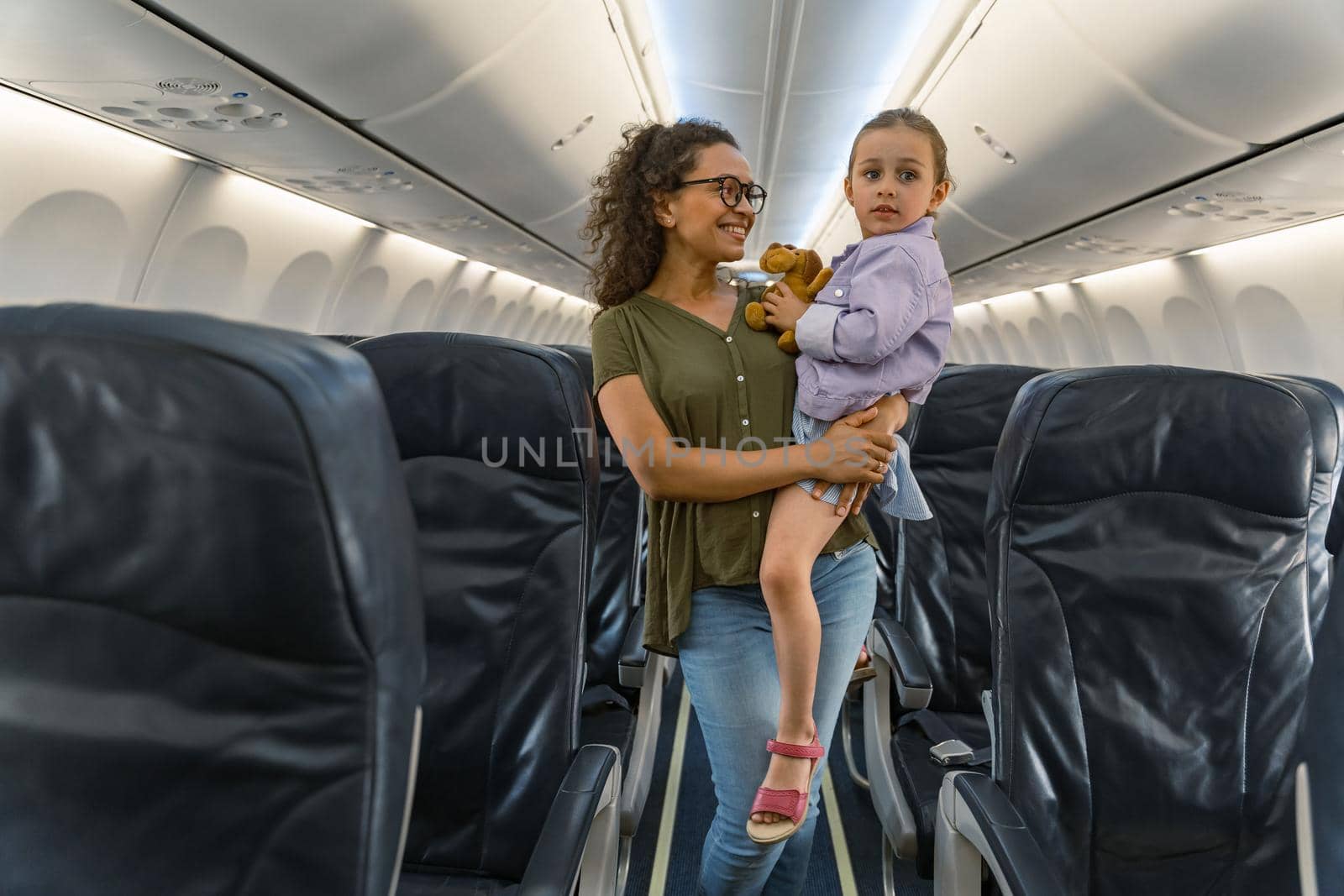 Smiling pretty woman holding kid while standing in airplane before the flight. Trip concept