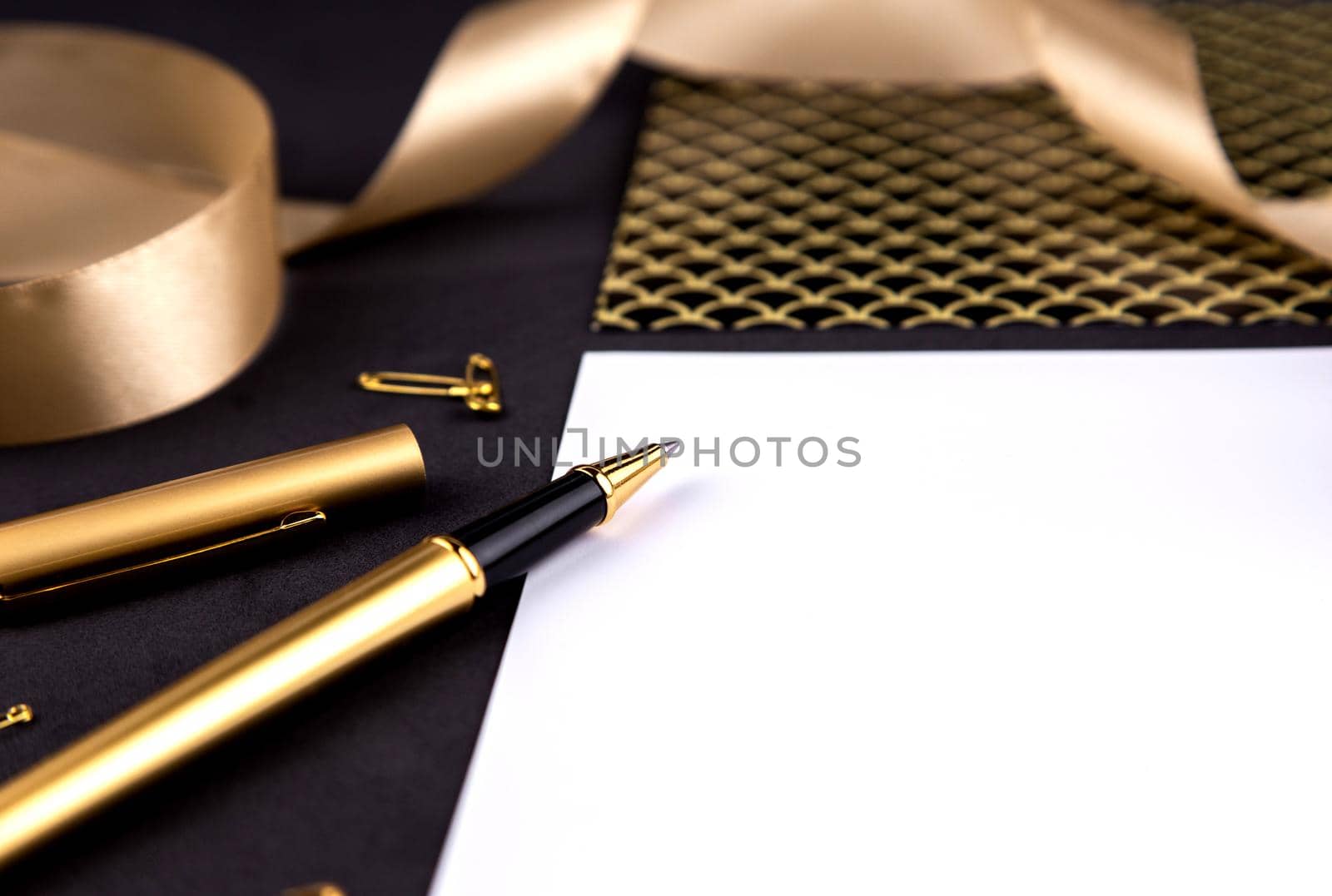 Gold pen, ribbon, paper clips and stationery on a black background with a white sheet of paper with copy space by lunarts