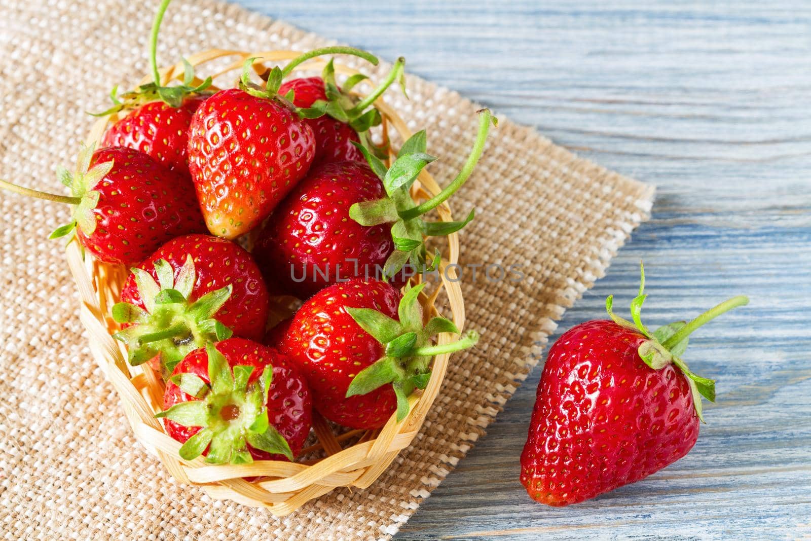 Fresh ripe strawberries in the basket and on burlap on the blue wooden background. Selective focus.