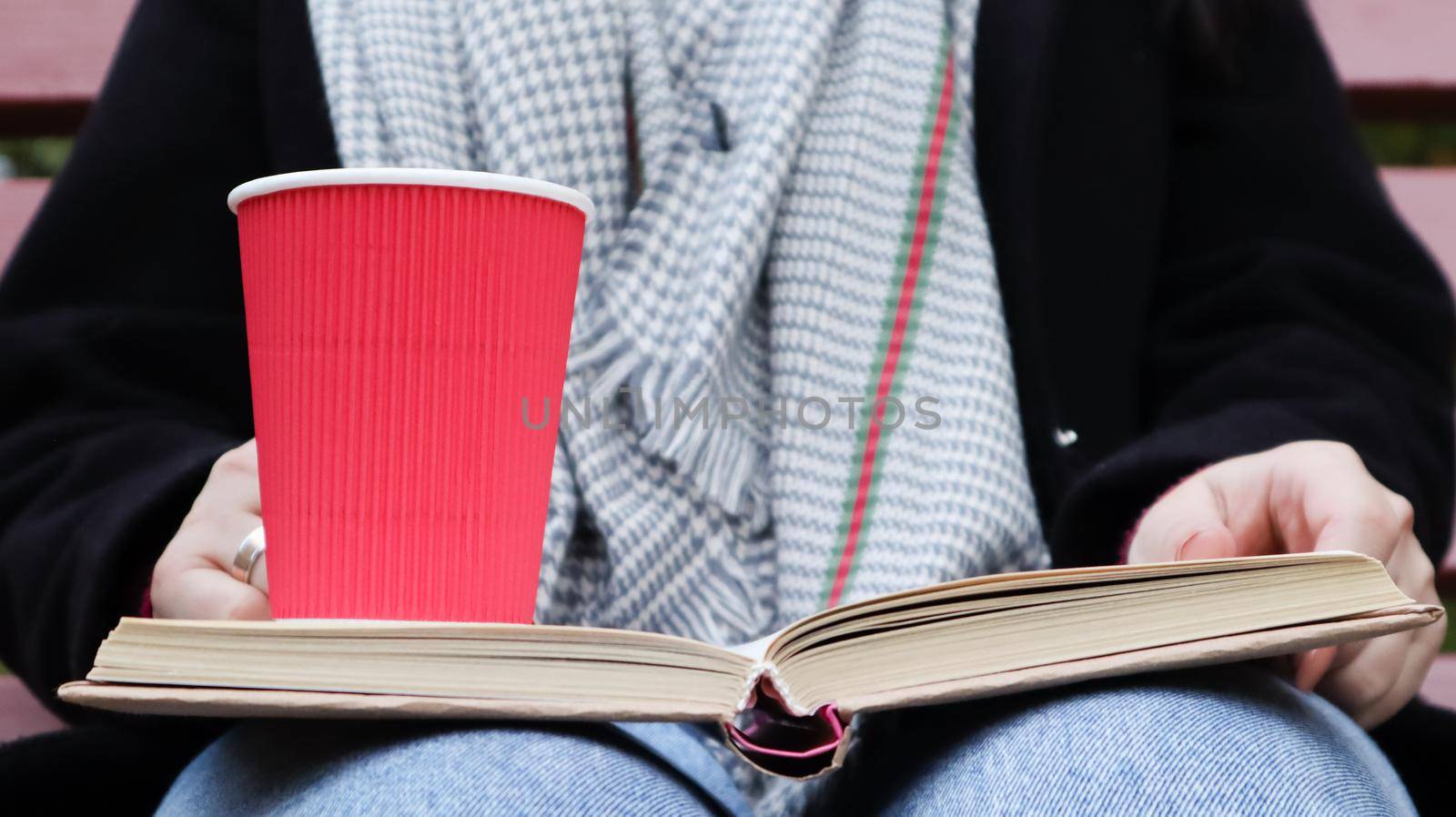 Young woman in jeans, coat and scarf, on a park bench. A woman is reading a book and drinking coffee or other hot drink outdoors alone. Close-up. The concept of honor, study, leisure and recreation