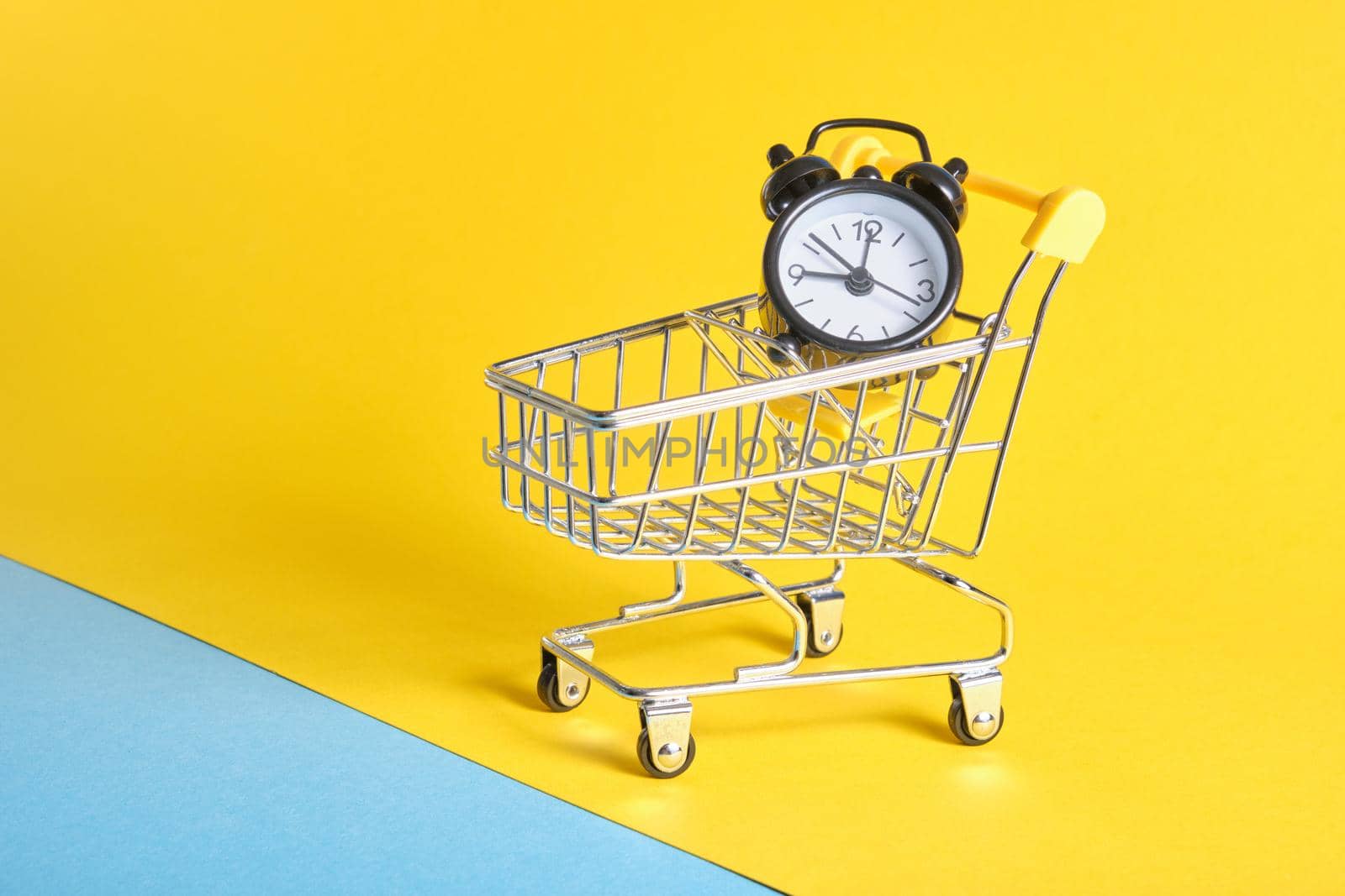 alarm clock in miniature shopping trolley on yellow background copy space