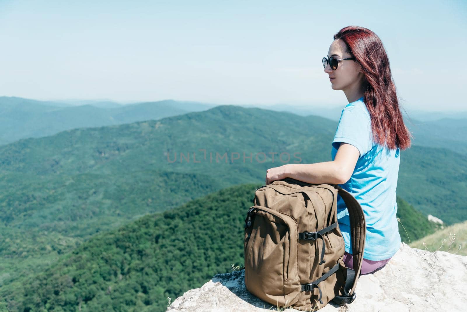 Hiker young woman with backpack sitting on edge of cliff high in summer mountains outdoor.