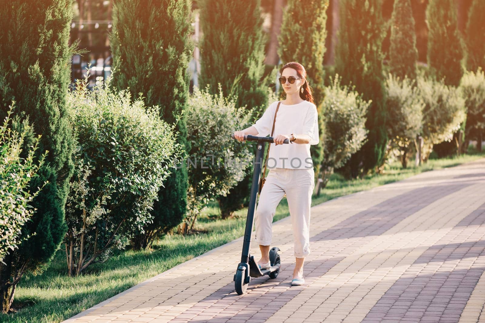 City stylish beautiful young woman riding an electric scooter in summer park.