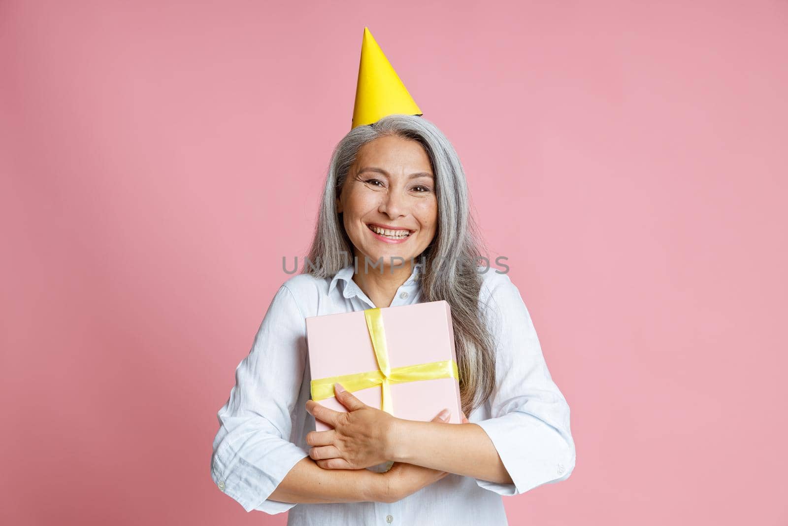 Joyful middle aged Asian lady with long hoary hair and bright party hat holds gift box standing on pink background in studio