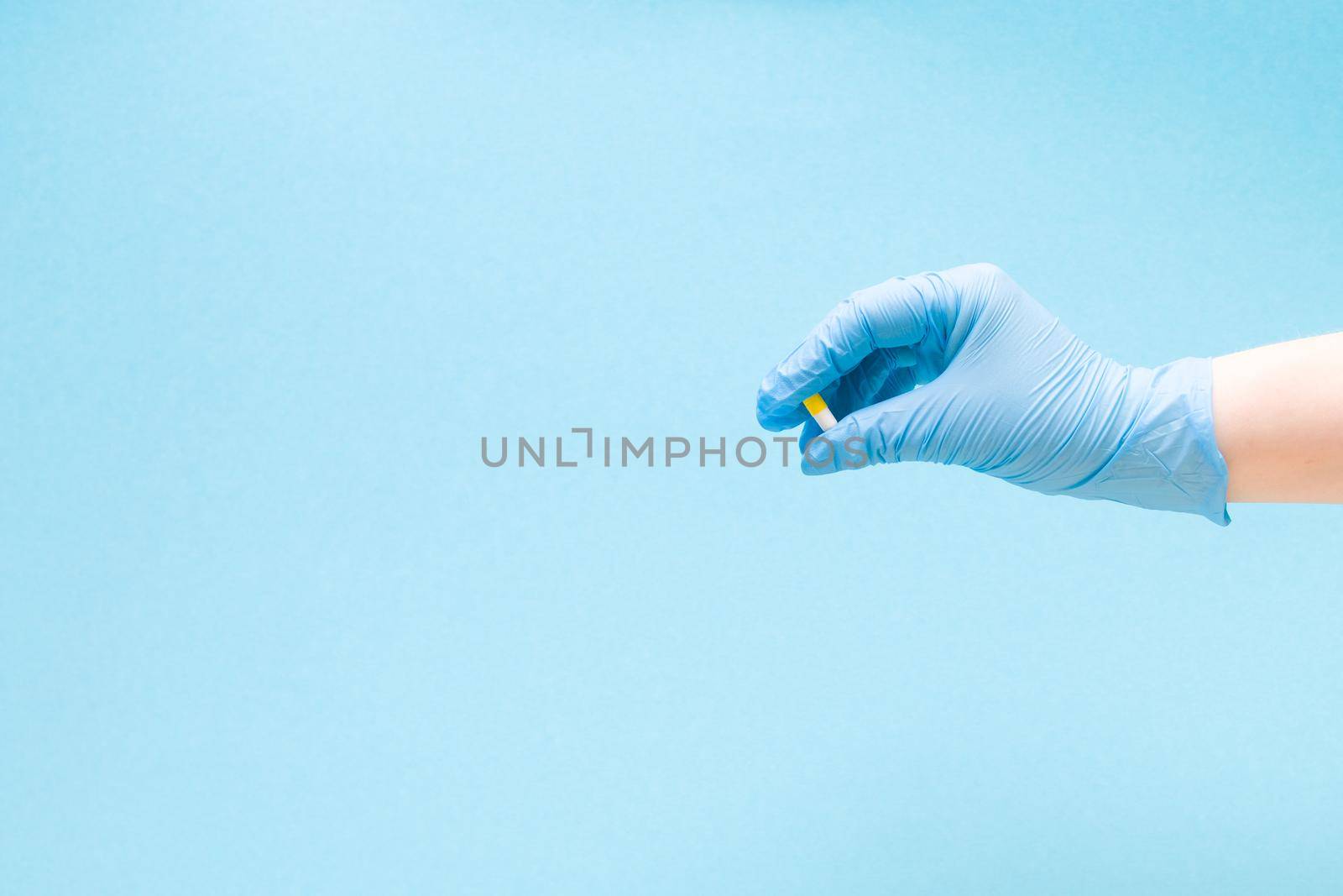 a female hand in a blue rubber medical glove holds a capsule tablet on a blue background copy space, tablet of white and yellow color, treatment and health care concept by natashko