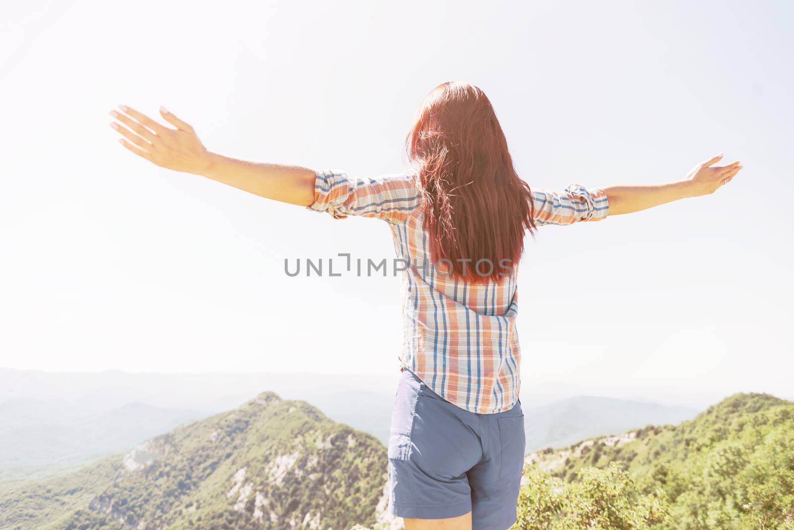 Rear view of freedom happy explorer young woman standing with raised arms high in mountains on sunny summer day.