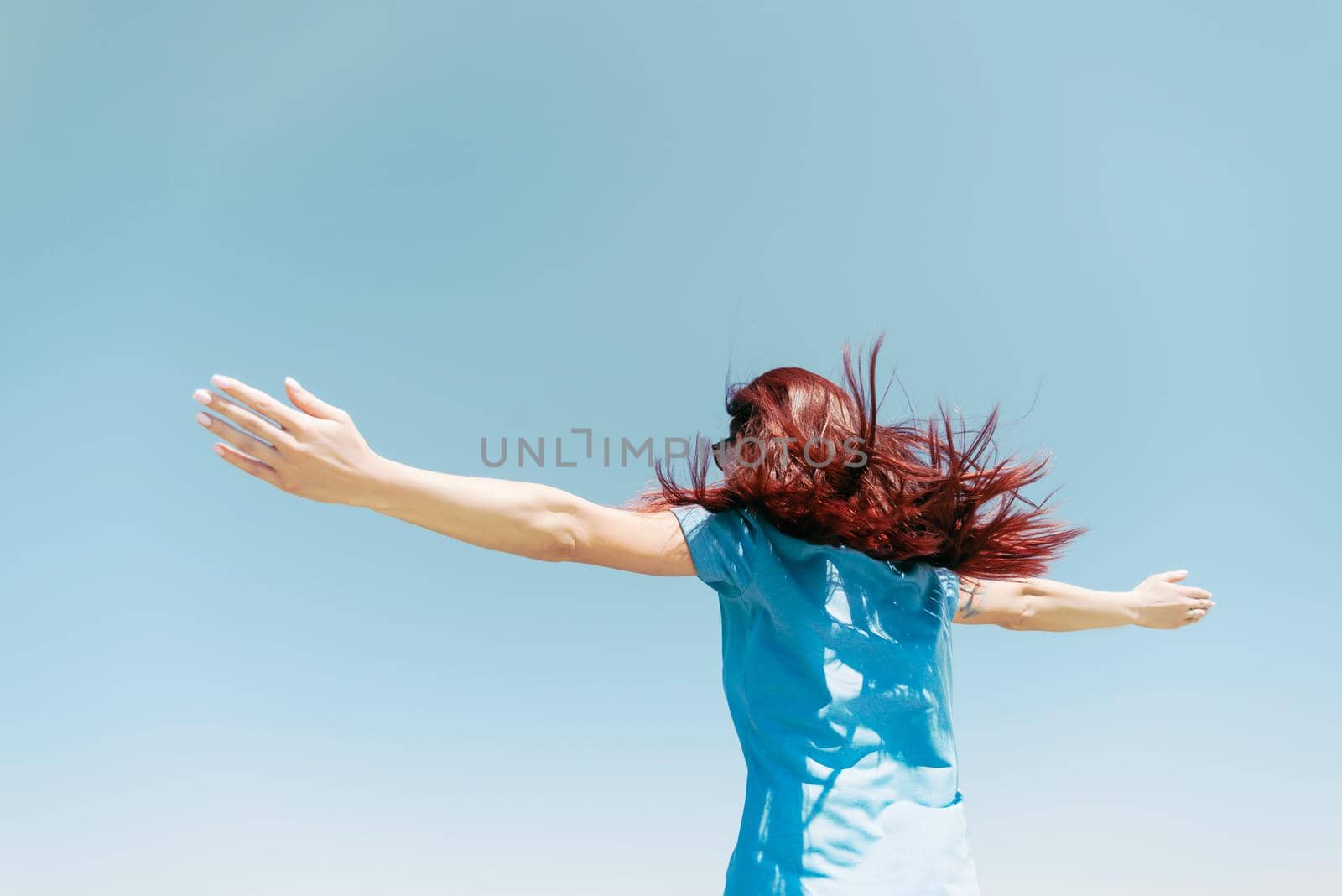Happy young woman with fluttering long hair standing with raised arms on background of blue sky.