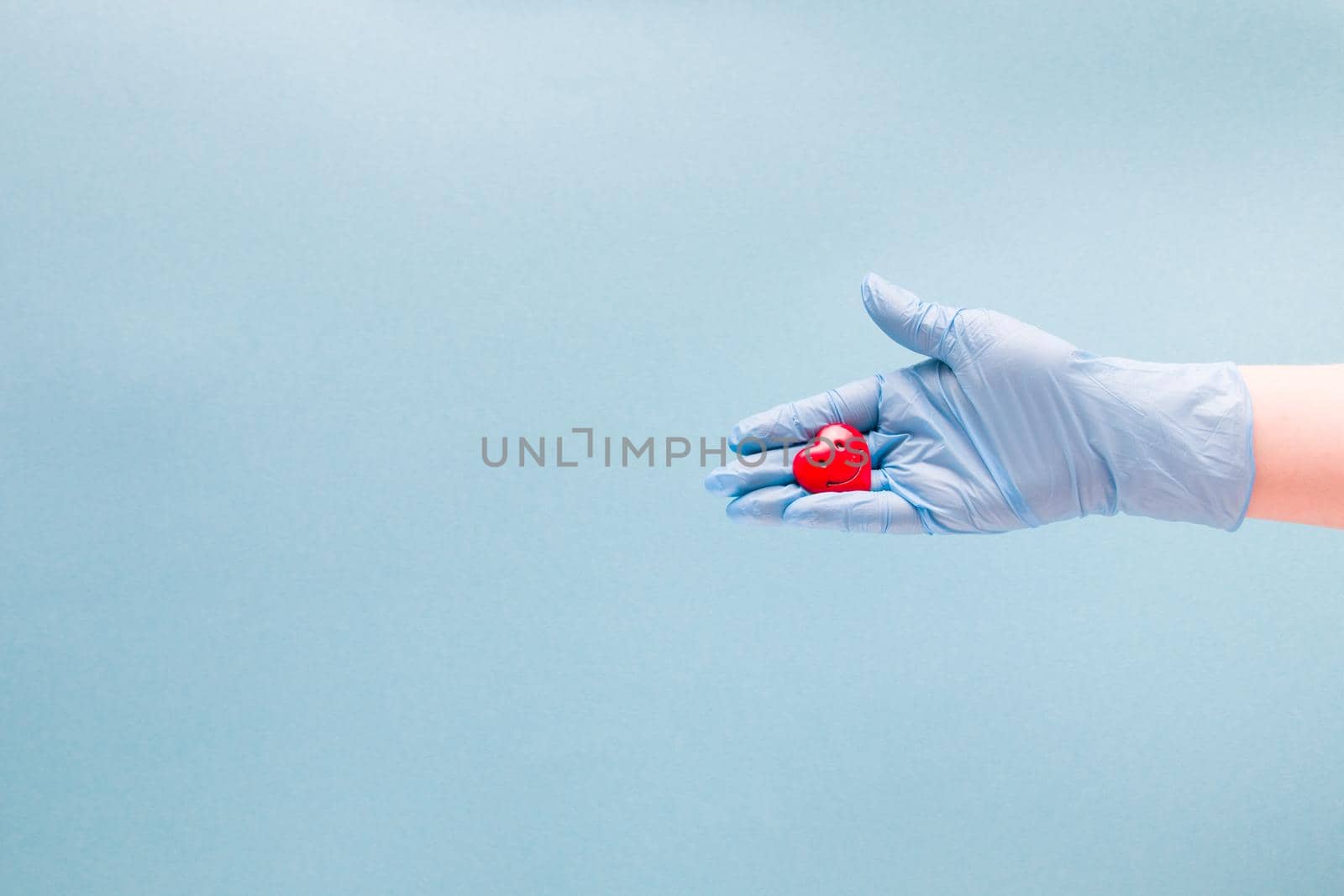 a female hand in a blue disposable medical glove holds a small red heart in her palm with a smile, a smiling heart, health care concept, blue background, copy space, varnish cordiologist cares about heart health