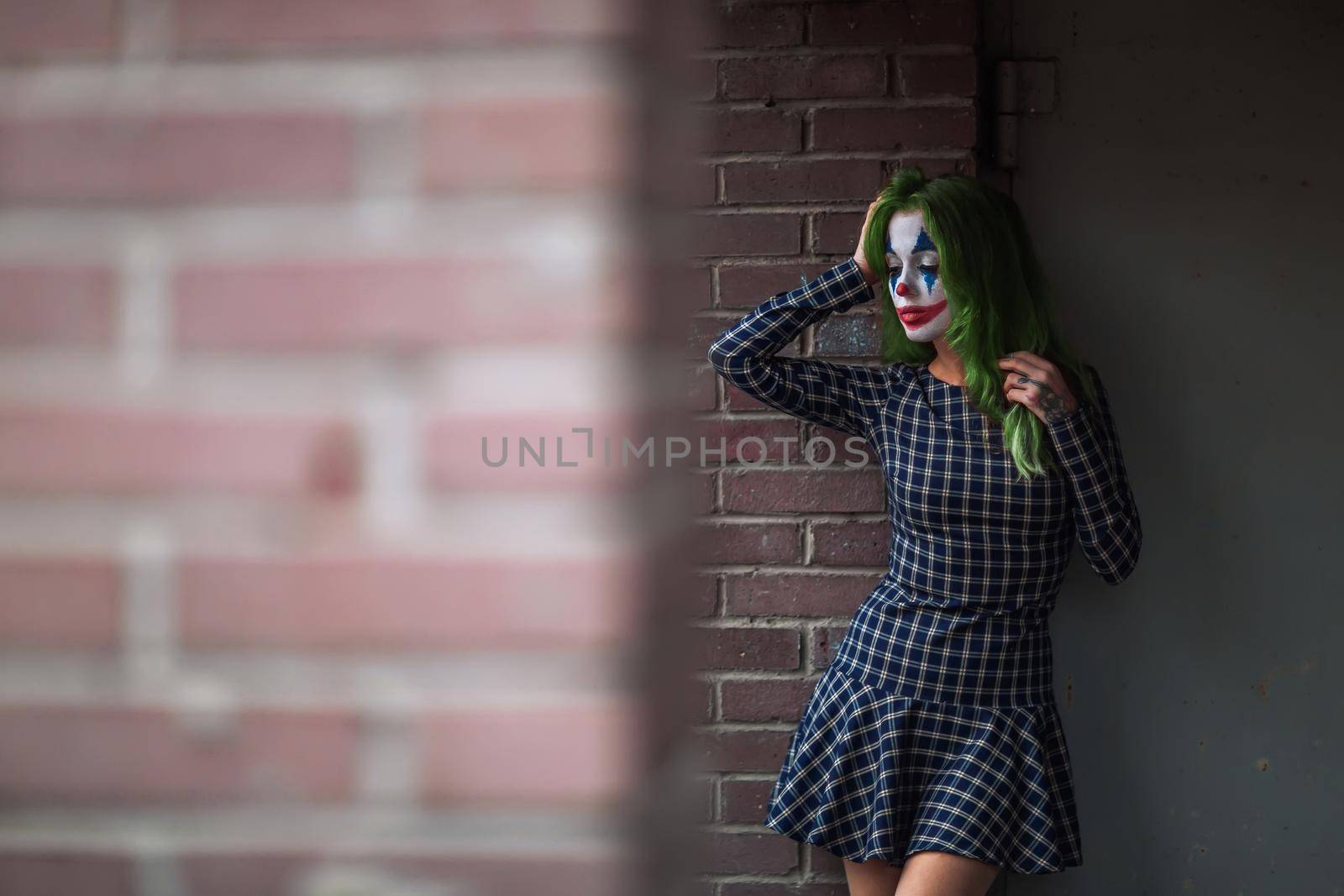 Portrait of a greenhaired girl in chekered dress with joker makeup on a brick wall blurred background.