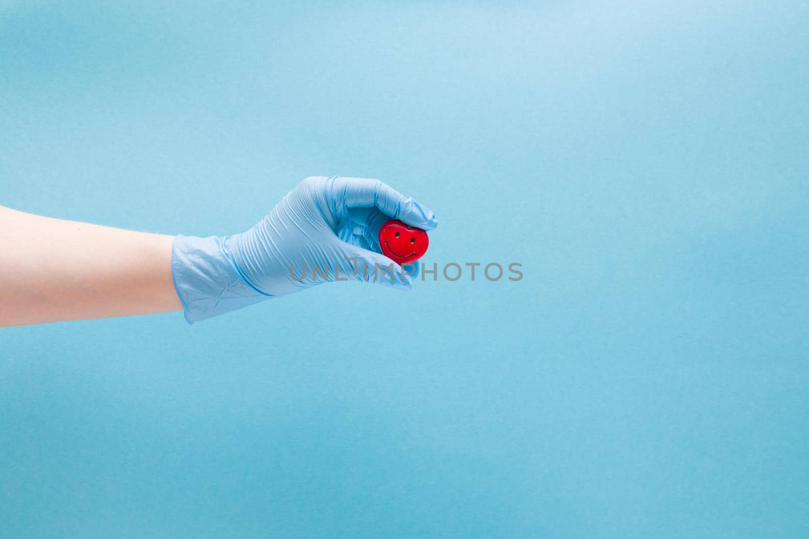 female hand in blue disposable medical glove holds a small red heart with a smile, smiling heart, health care concept, blue background, copy space, vari cordiologist treats