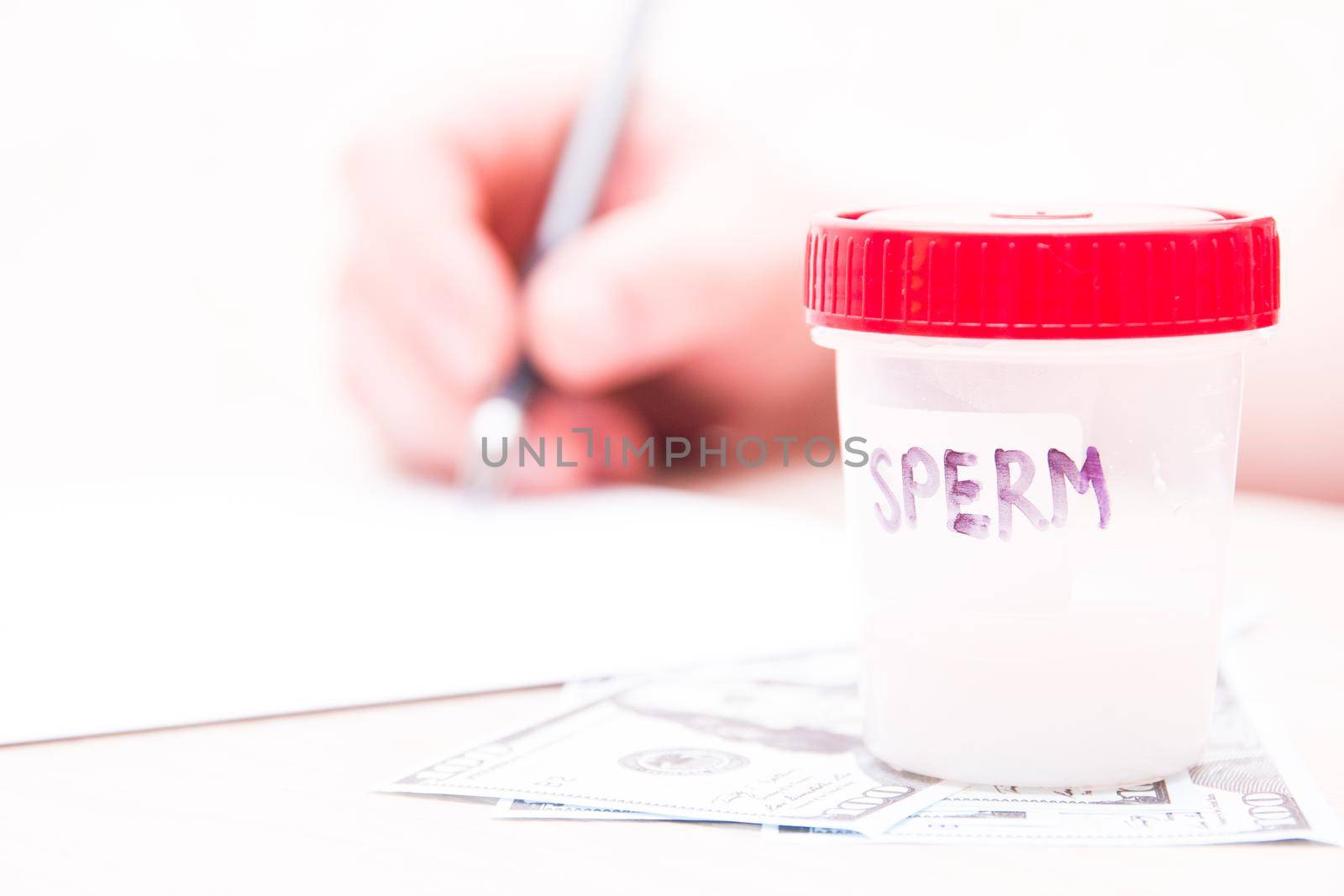 a test jar with a red cap and the inscription â Spermâ  stands on dollar bills, against the background a man signs documents, a copy place, sperm in a jar, a sperm donor concept, sell sperm, close up