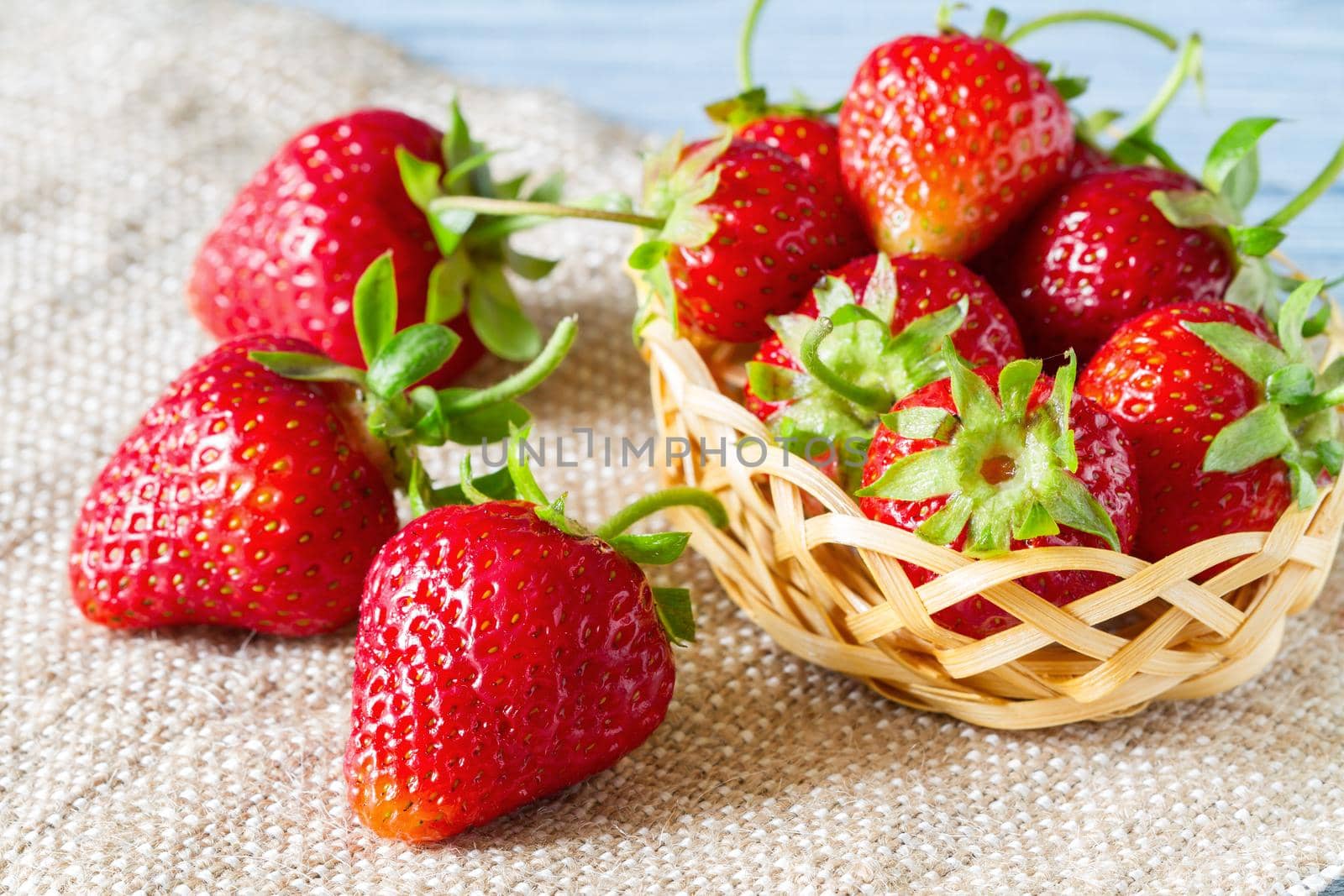 Fresh ripe strawberries in the basket and on burlap on the blue wooden background. Selective focus.