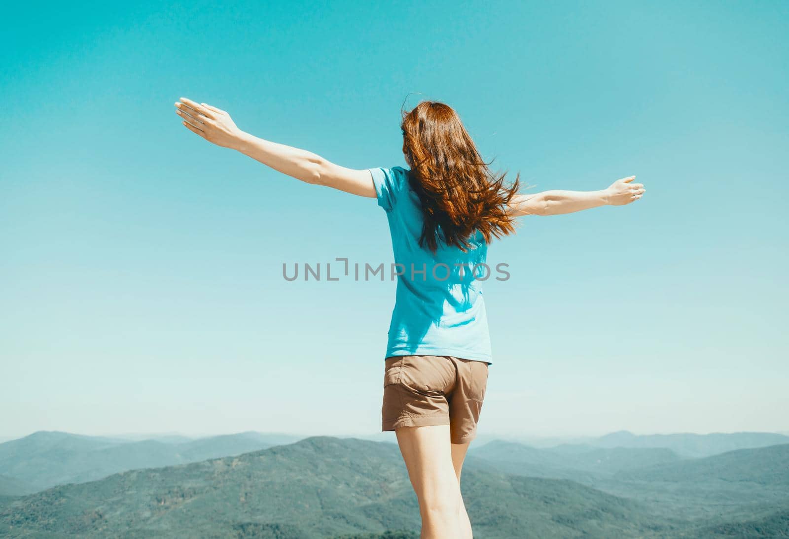 Happy young woman standing with raised arms on background of blue sky and green mountains in summer outdoor, rear view.