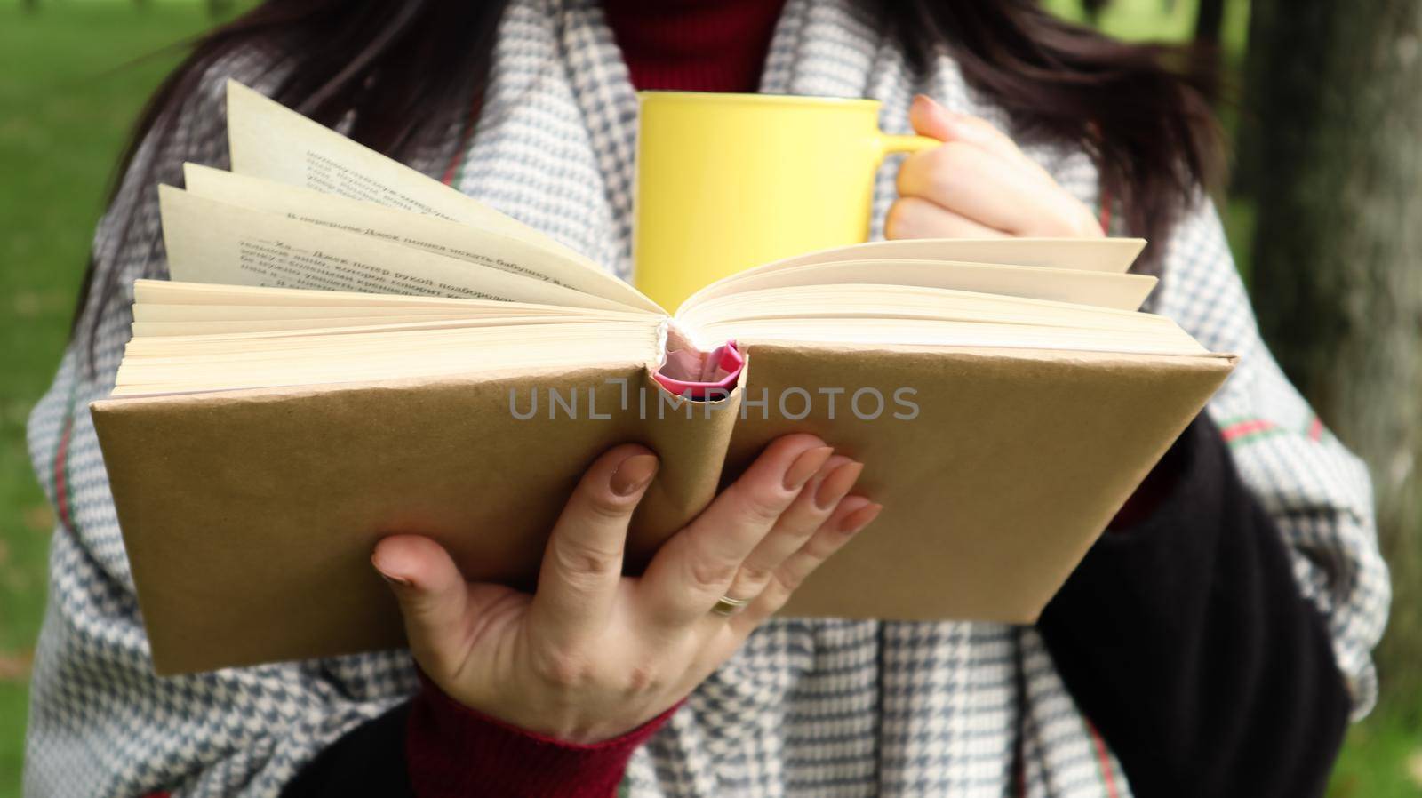 A girl dressed in a coat and a scarf in the autumn forest holds a book and a cup with a hot drink in her hands close-up in a city park on a warm day. The concept of reading, relaxation and comfort. by Roshchyn