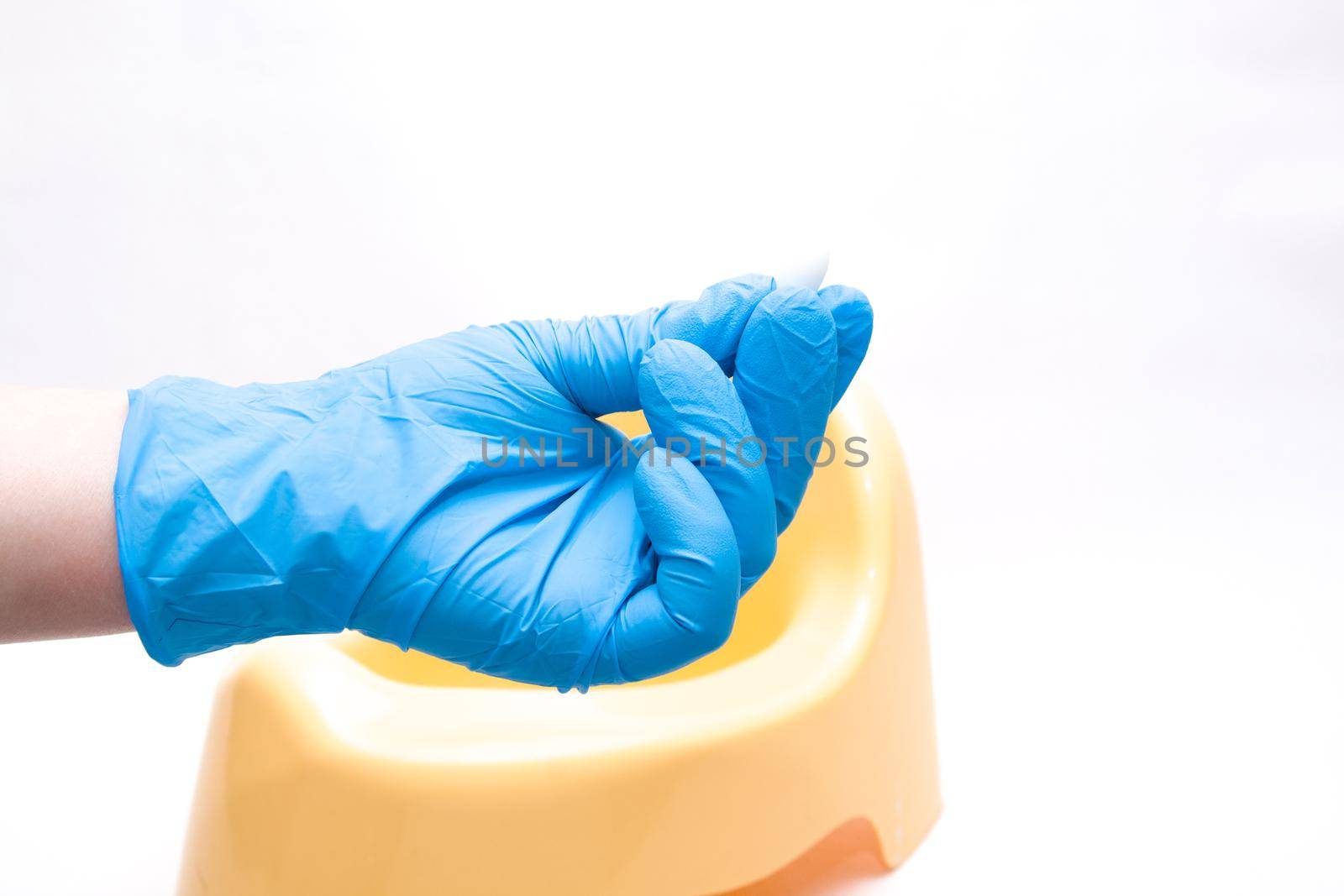 a hand in a disposable blue medical glove holds a test jar with a white spoon for collecting feces, in the background is a yellow children's pot, white background, copy space by natashko