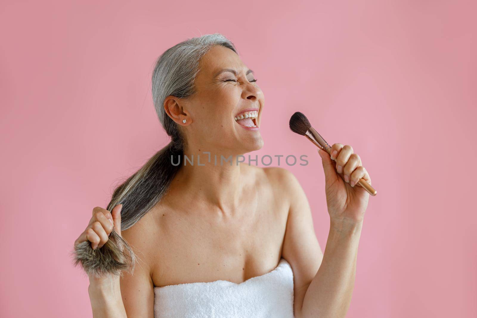 Jouful middle aged Asian woman sings song using cosmetic brush as microphone on pink background in studio. Mature beauty lifestyle