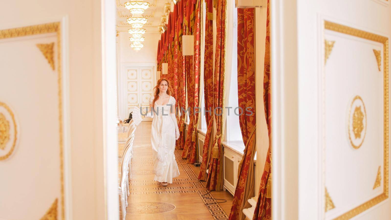 Red-haired woman in vintage ball costume is walking in the ball room, telephoto shot