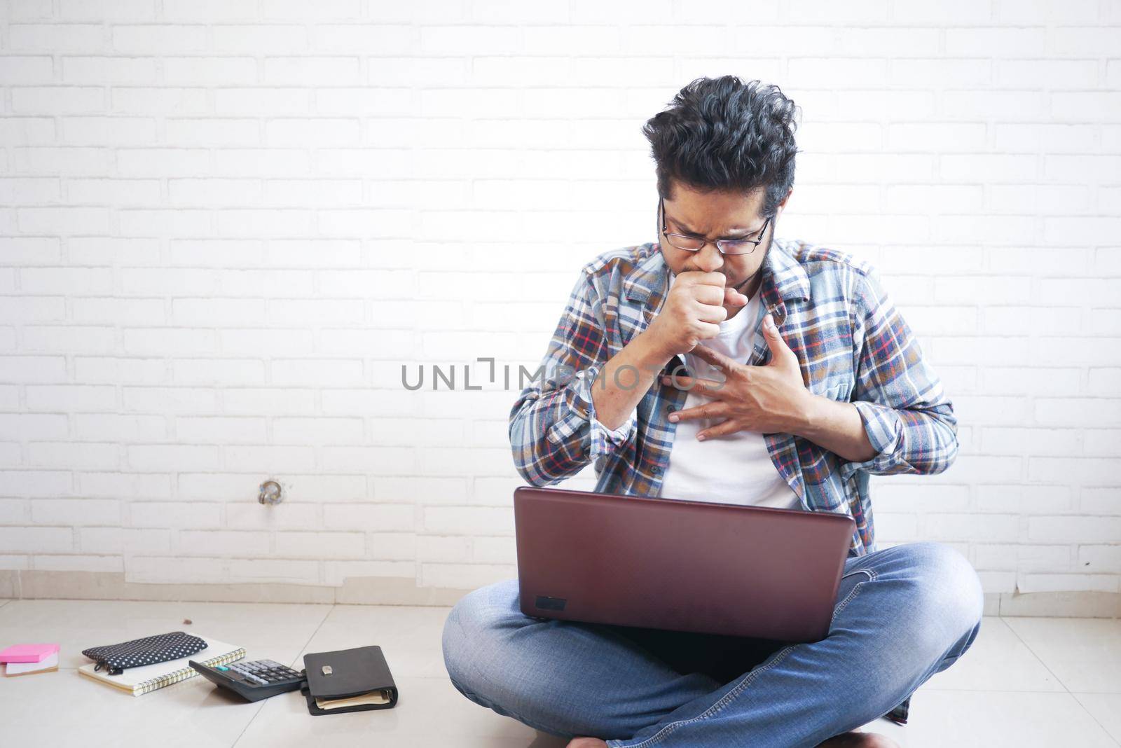 young sick man coughing while working on laptop sitting on floor by towfiq007